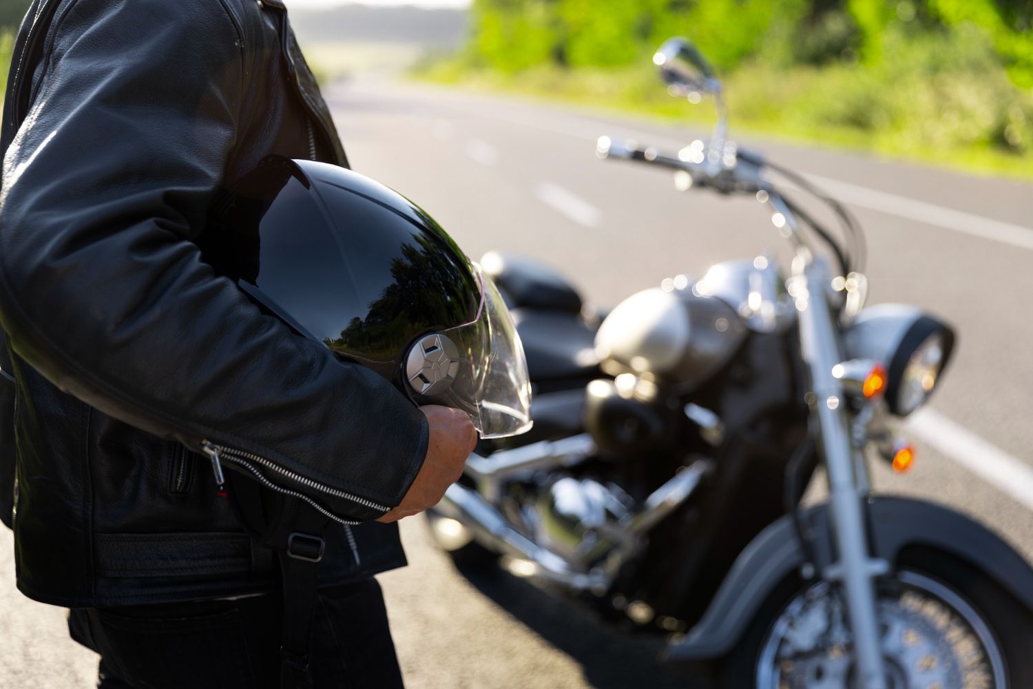 Person in black leather jacket holding a helmet next to a parked motorcycle on a road.