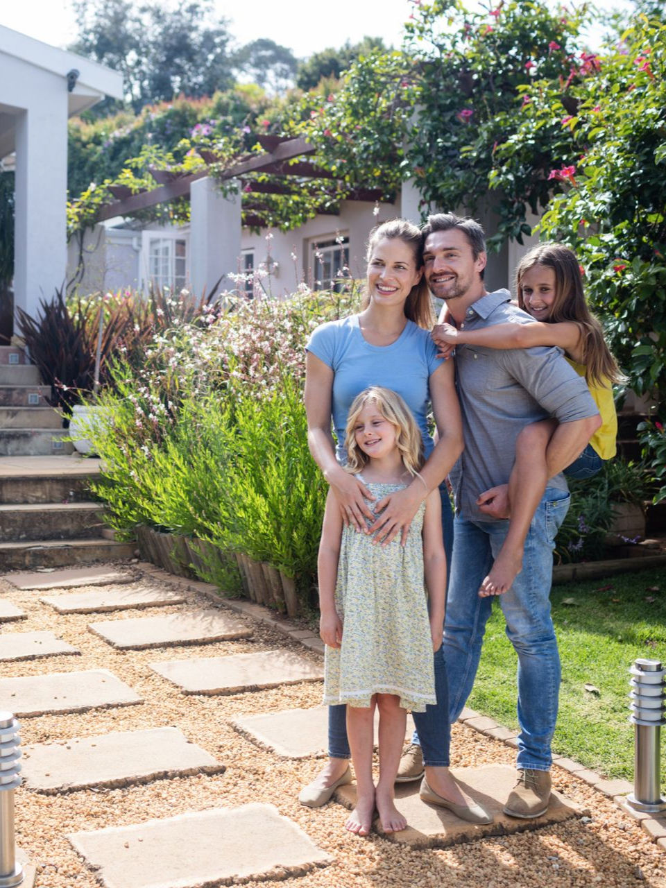 Family of four smiling outdoors in front of a house. Stone path, green plants, sunny day.
