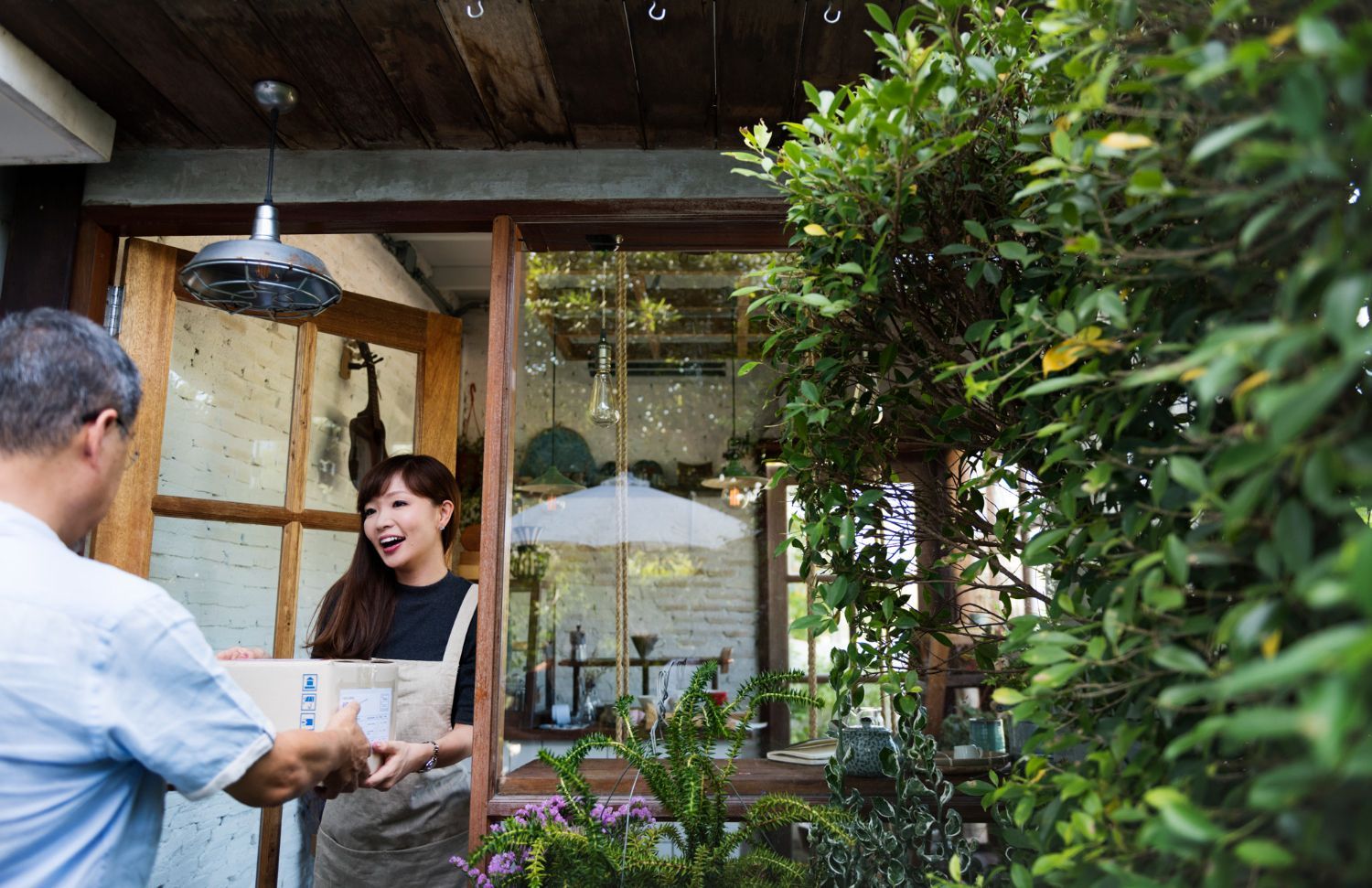 Man handing a package to a woman at a shop entrance. Woman smiles, greenery surrounds.