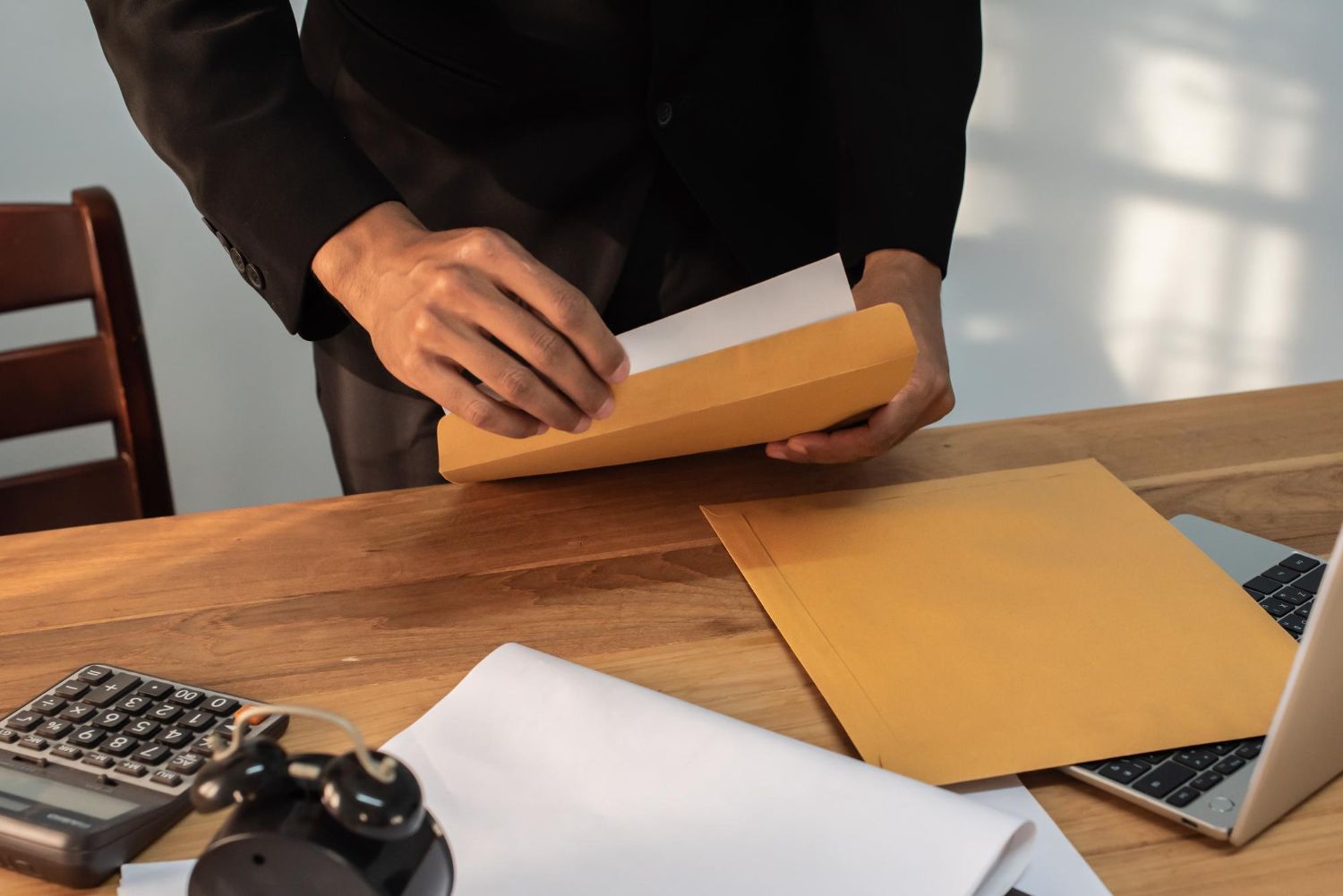 Person placing paper into a brown envelope at a wooden desk with a calculator, laptop, and clock.