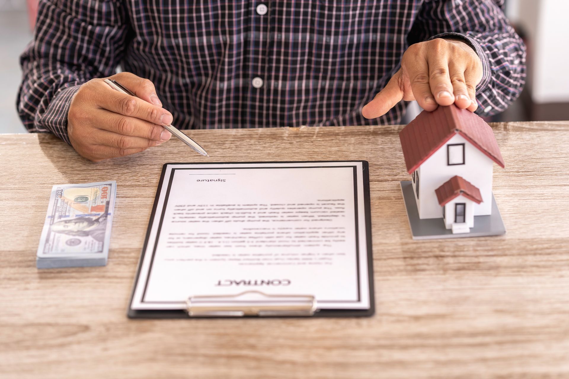 Person with pen pointing at a contract on a desk, next to a house model and cash.