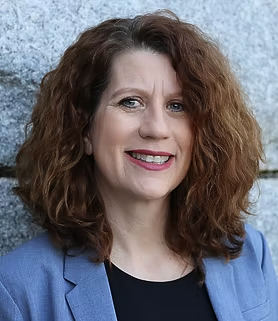Woman with curly auburn hair smiles, wearing a black top and light blue blazer, leaning against a gray stone wall.