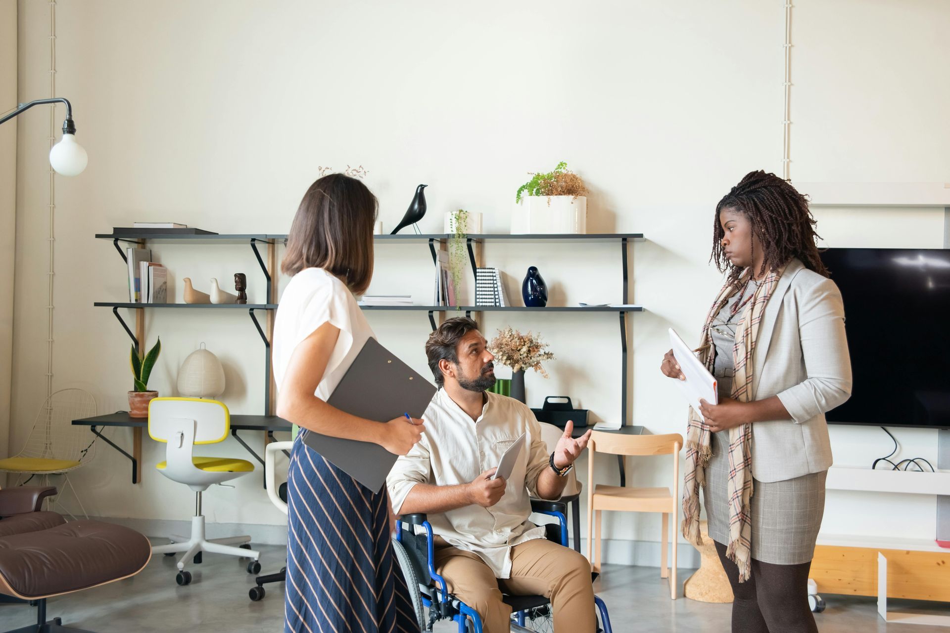 A man in a wheelchair is talking to two women in a living room.