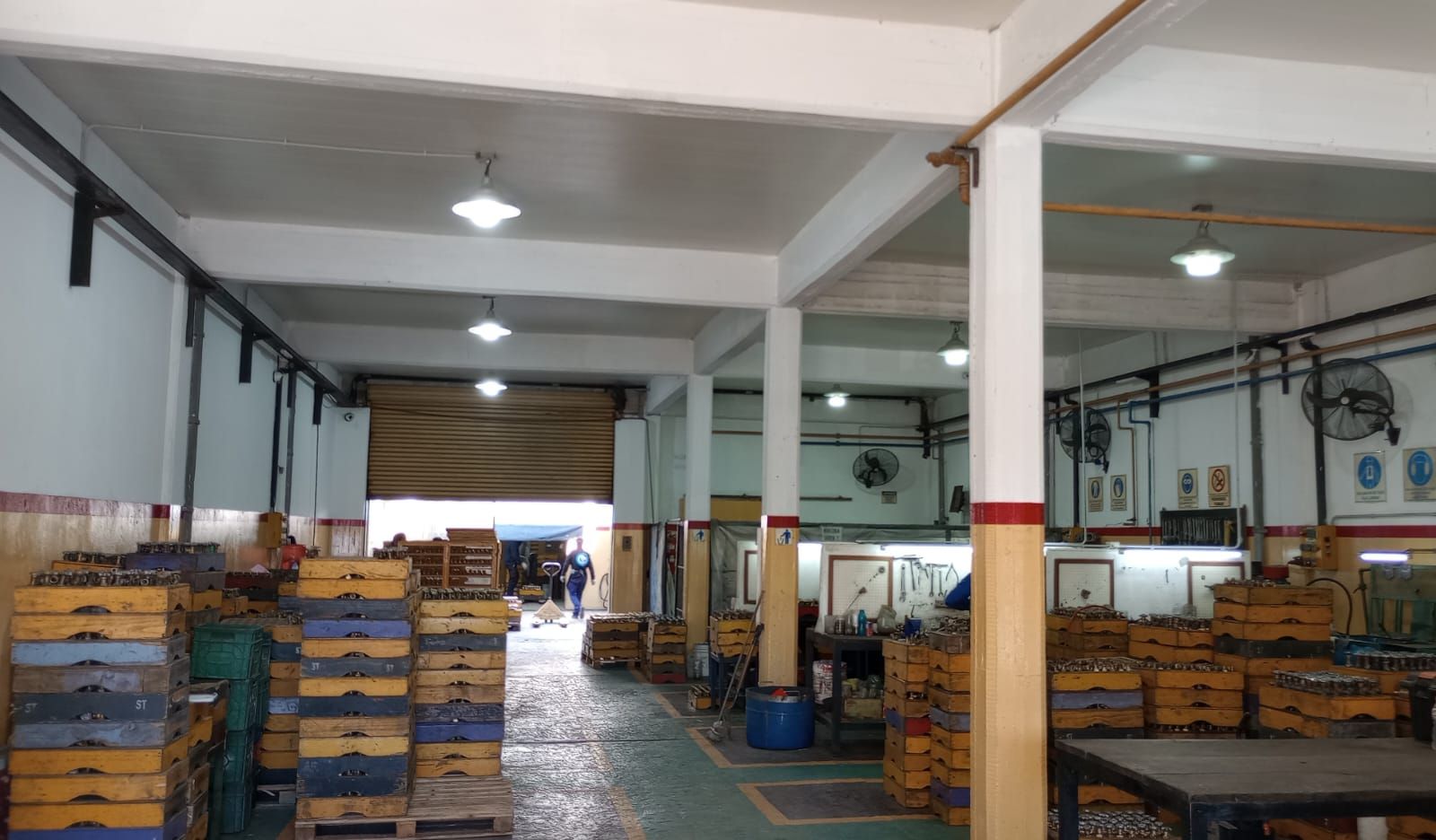 Dentro de un almacén: cajas apiladas, puerta abierta, luces en el techo, paredes blancas con detalles en rojo.