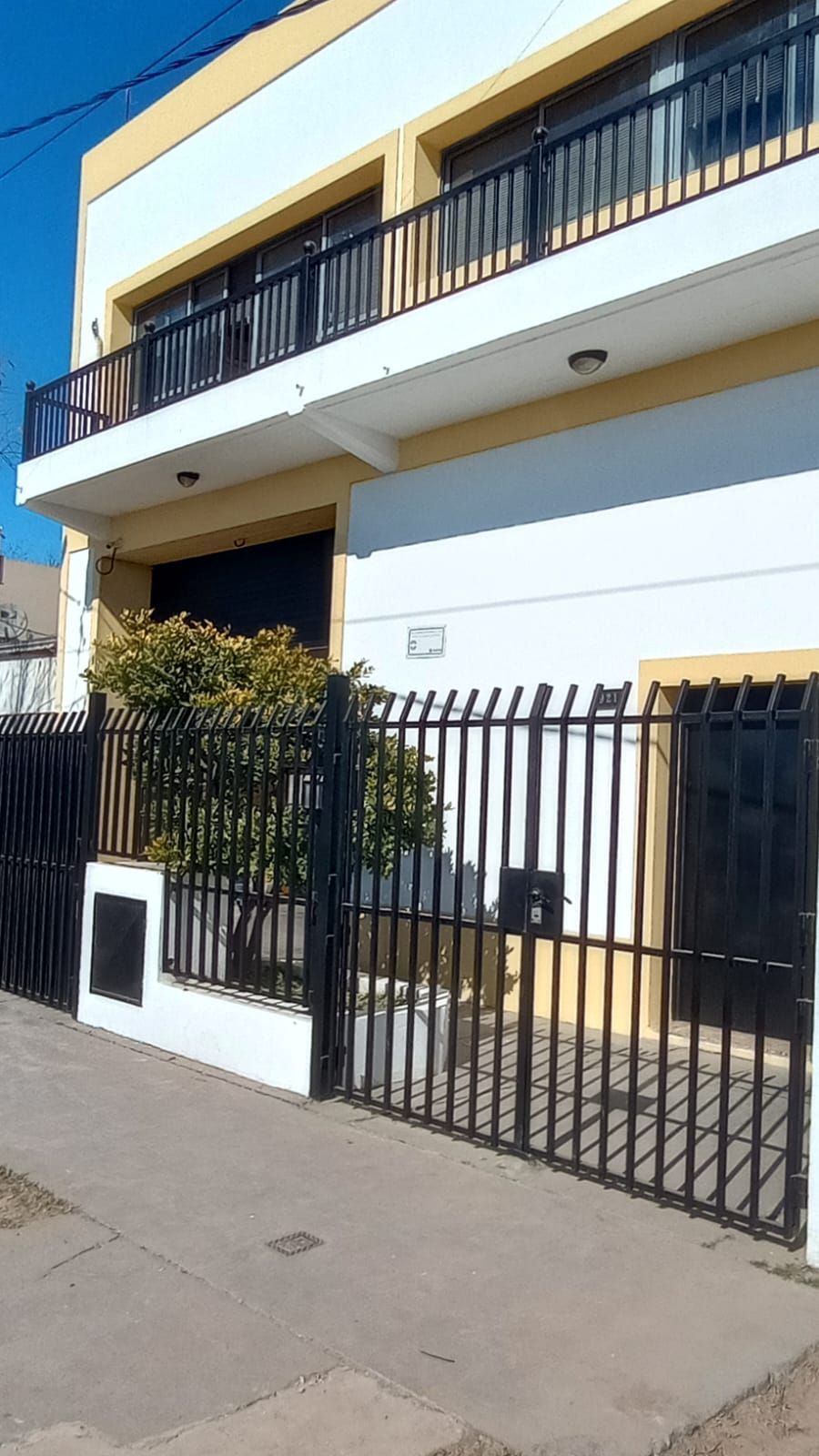 Edificio de dos plantas con barandillas negras, portón y un pequeño árbol. Exterior blanco y amarillo con acera de hormigón.