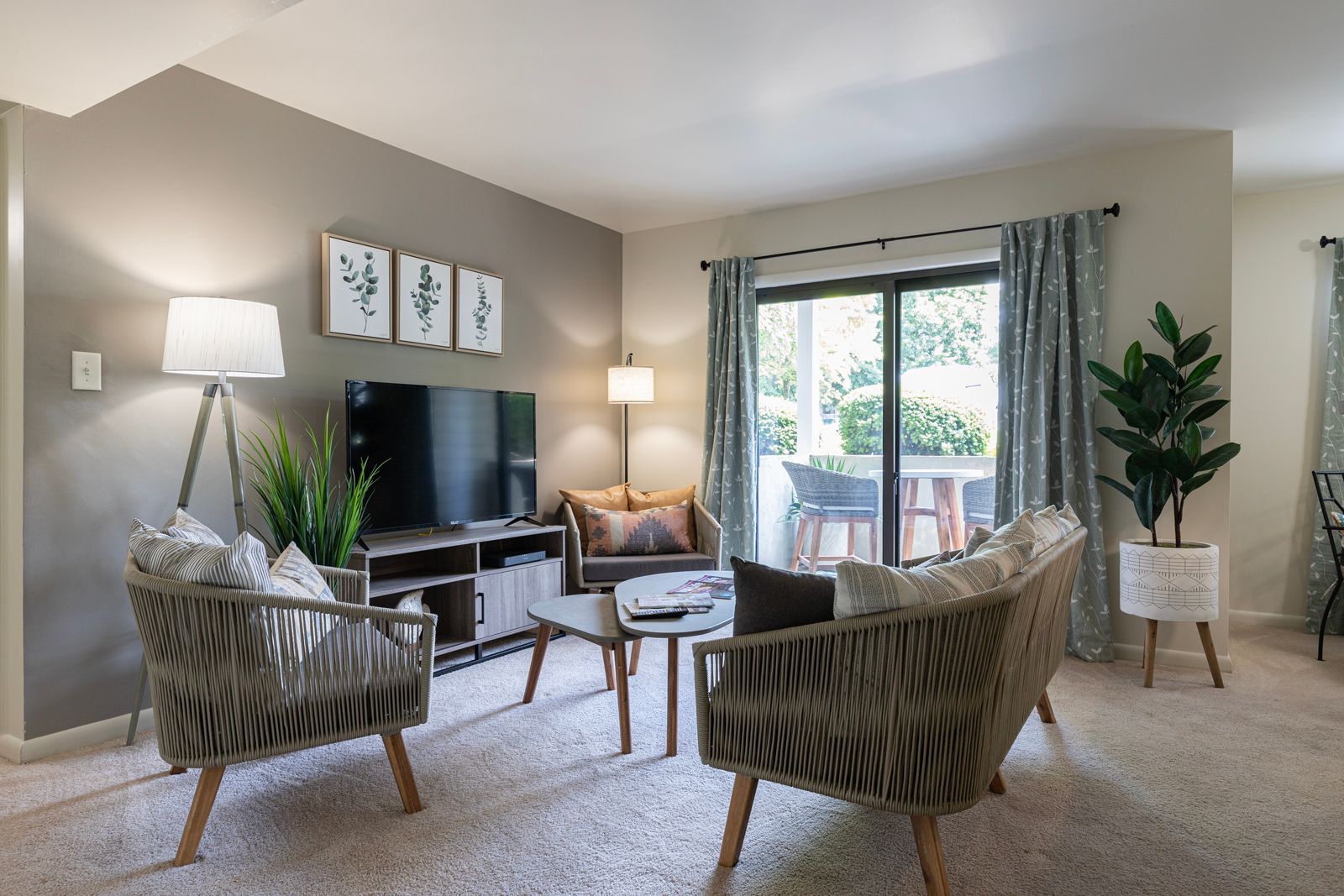 Bright, modern living room with two woven armchairs, a small coffee table, TV on a media console, and sliding glass doors leading to a patio.