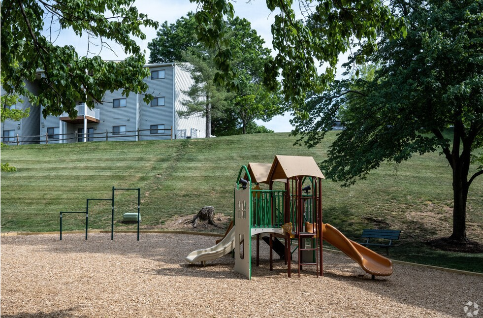 Playground with slide and climbing structures on wood chips; apartment building in the background on a grassy hill.