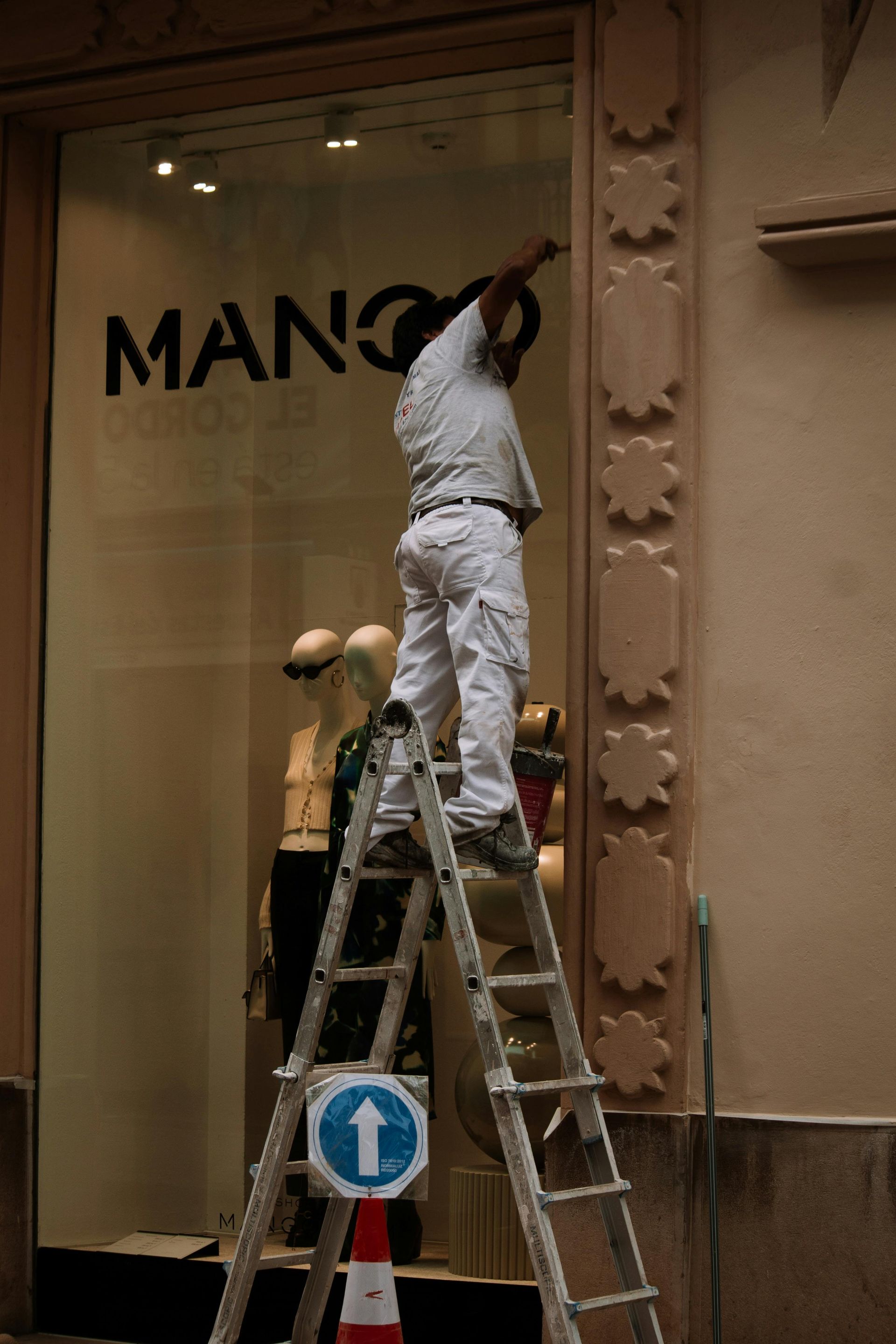Man on ladder adjusts sign at a Mango store window. Building exterior with mannequins and a street sign.