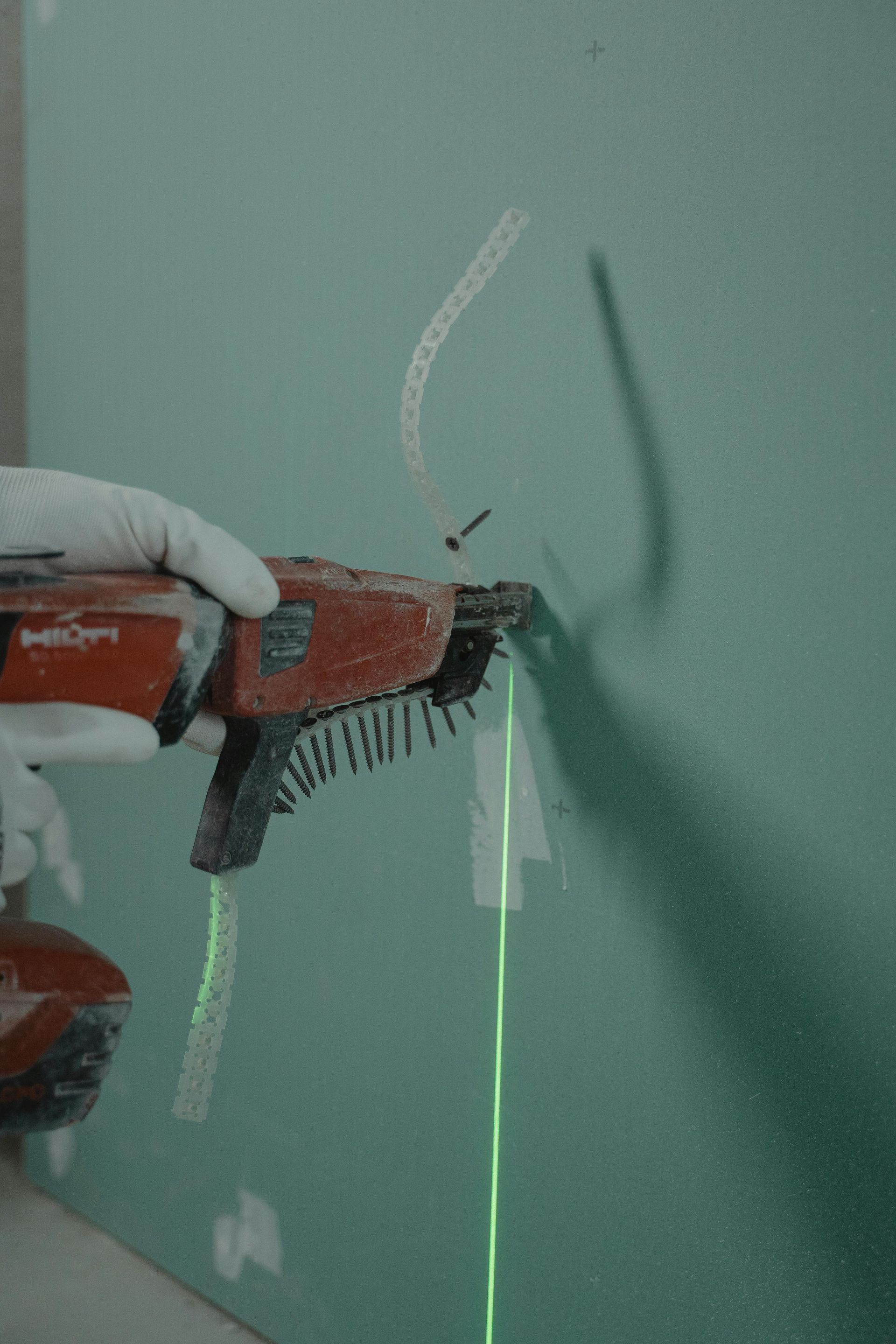 Person using a drywall screw gun to attach a metal strip to a green wall, guided by a laser level.