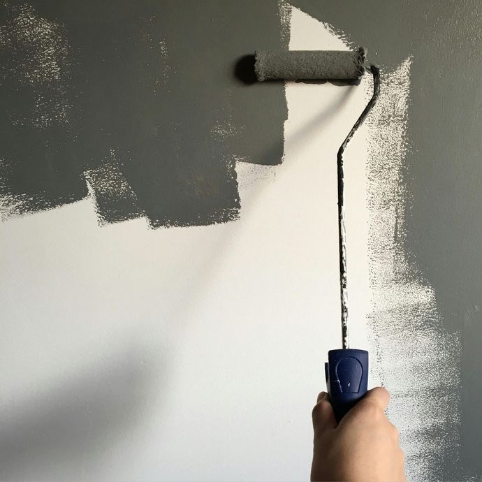 Hand holding a paint roller, applying gray paint to a white wall.