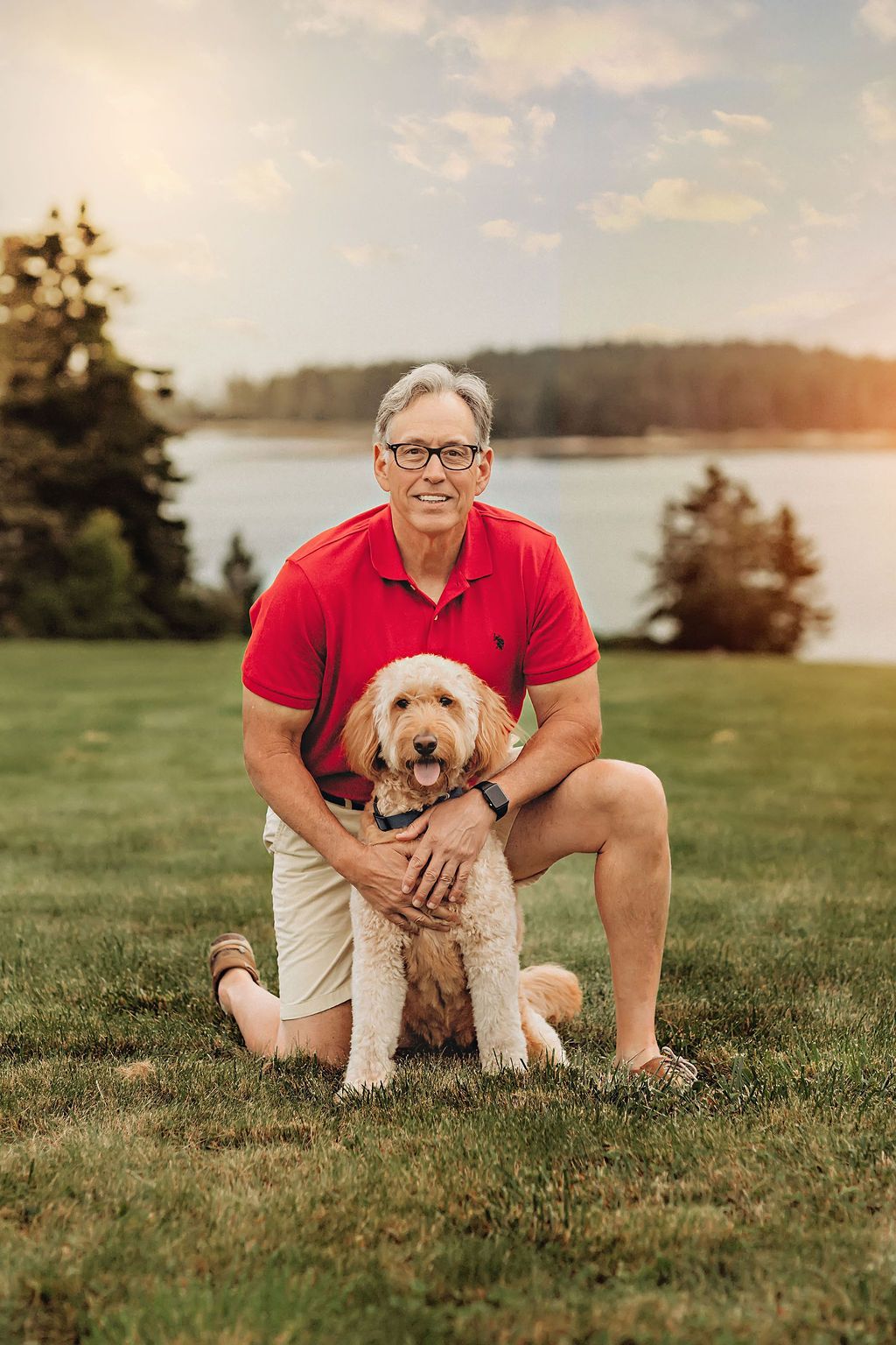 Man in red shirt and khaki shorts kneels, hugging a tan dog on grassy lawn; water and trees in the background.