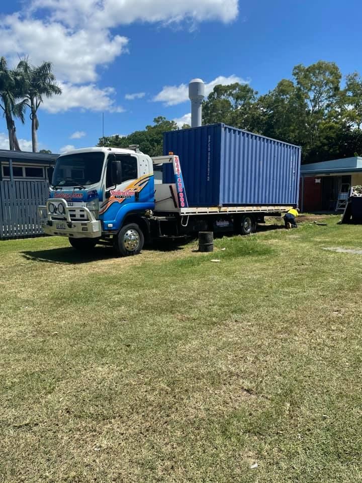 Blue shipping container on a flatbed truck parked on grass under a partly cloudy sky.