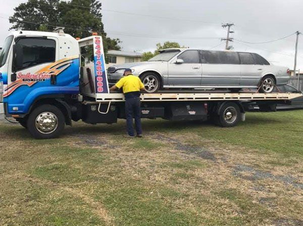 A Man is Standing Next to a Tow Truck With a Car on the Back — Southern Cross Tilt Trays in Andergrove, QLD
