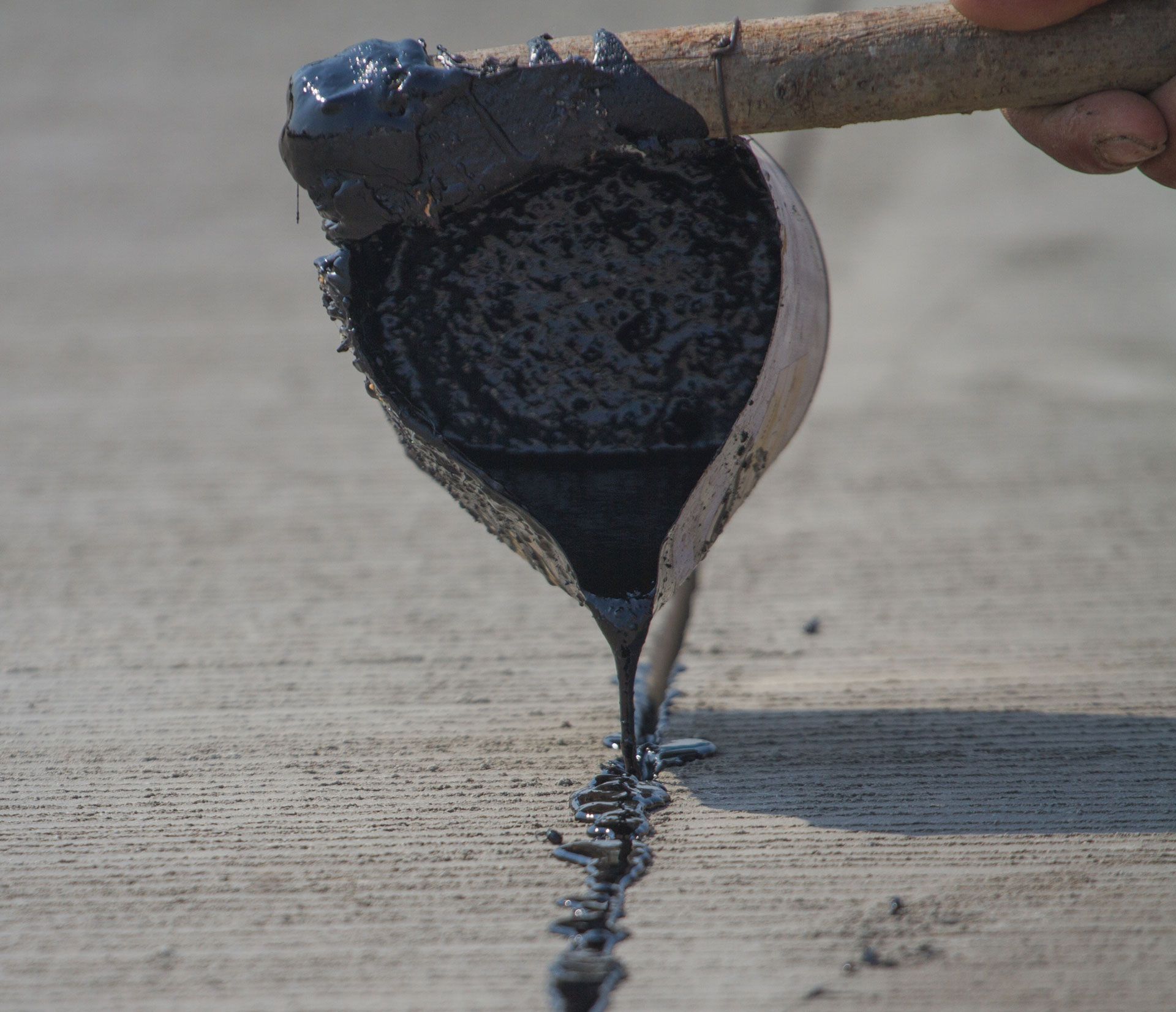 A person uses a handheld metal scoop to pour hot, black tar into a crack on a concrete road surface.