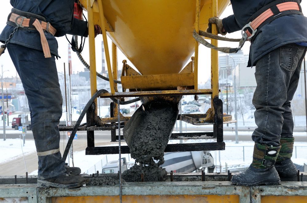 Two construction workers are pouring concrete into a container.