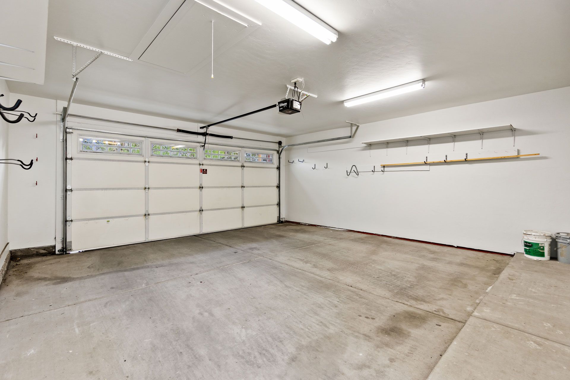 A clean, empty garage with white walls, a multi-paneled door, concrete floor, overhead lighting, and wall-mounted shelves.