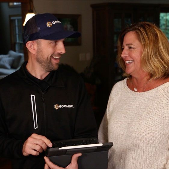 Man in black jacket with woman, both smiling, inside a home, man holding tablet.
