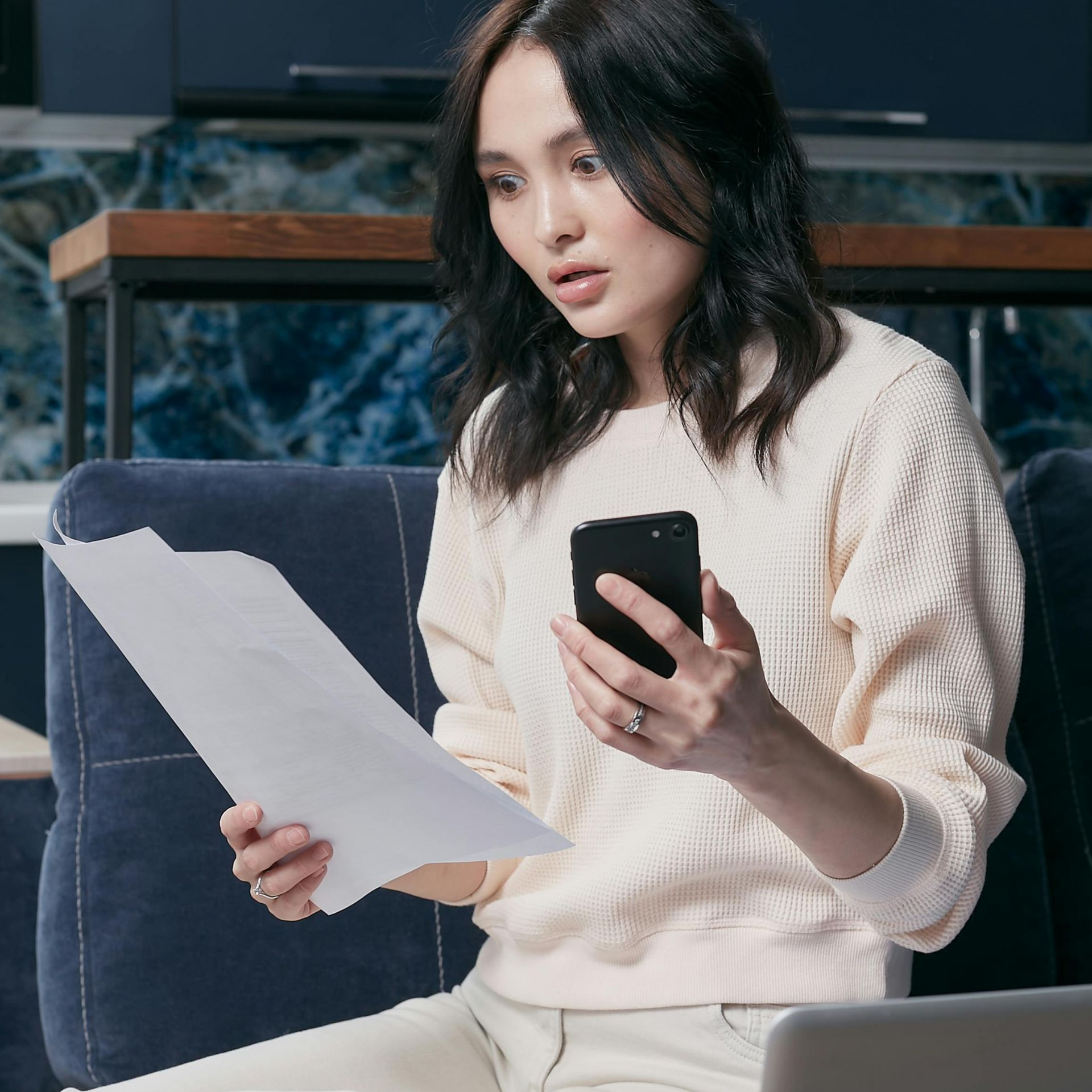 Woman on couch looking at phone and paper, laptop nearby.