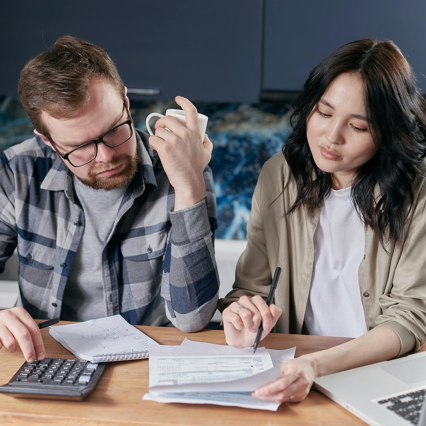 Couple reviewing finances at a table. Man uses calculator, woman holds pen, and laptop visible.
