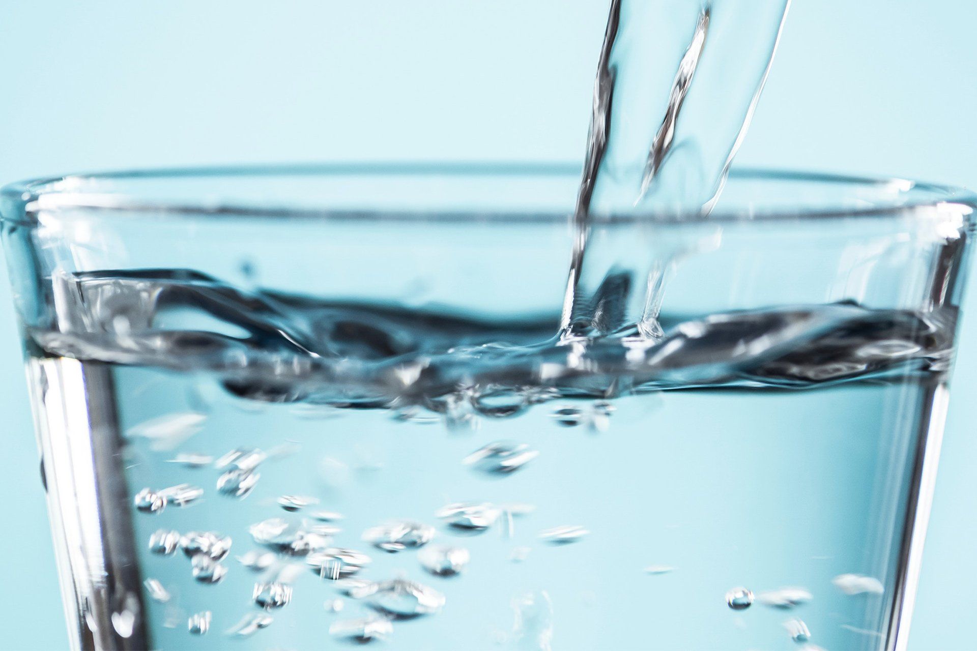 Water being poured into a clear glass, creating bubbles in the water, on a light blue background.