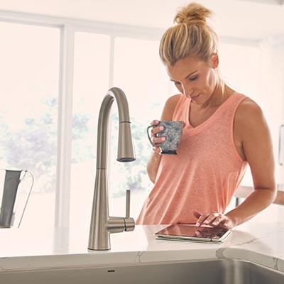 A woman is standing in front of a sink holding a cup of coffee and a tablet.