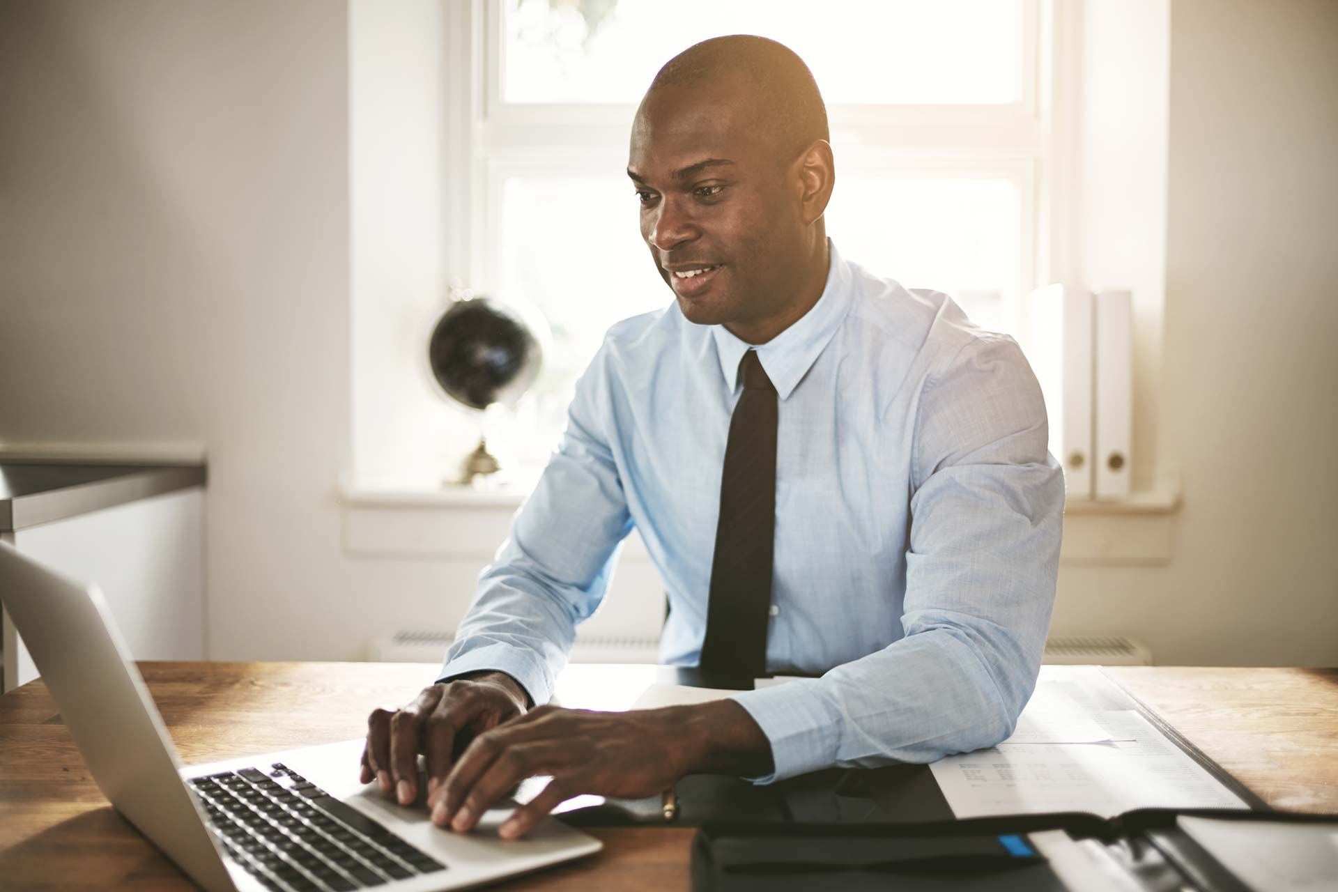 Man in a light blue shirt and tie working on a laptop at a desk in a well-lit office.