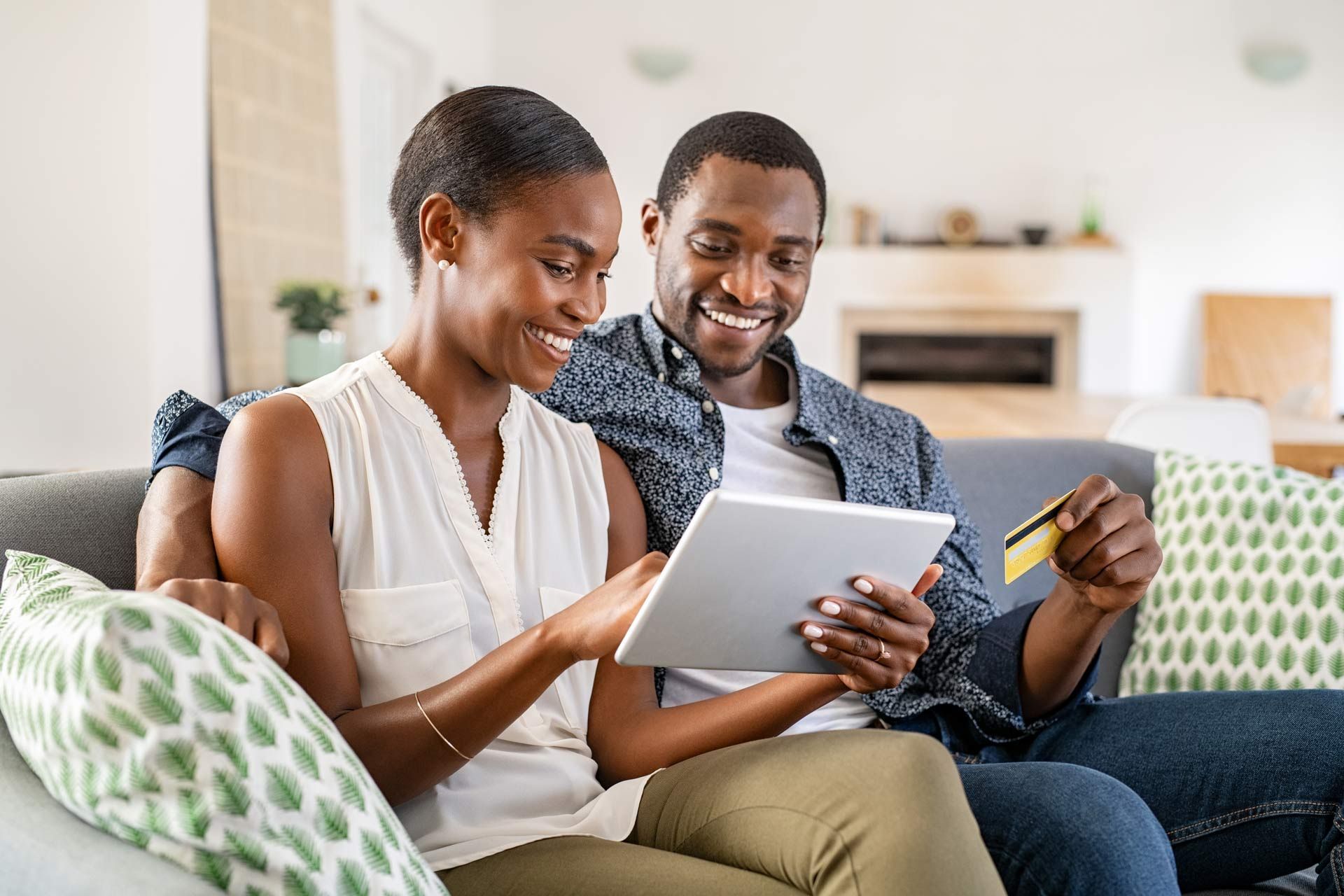 Couple on couch shopping online with a tablet and credit card, smiling in a living room.