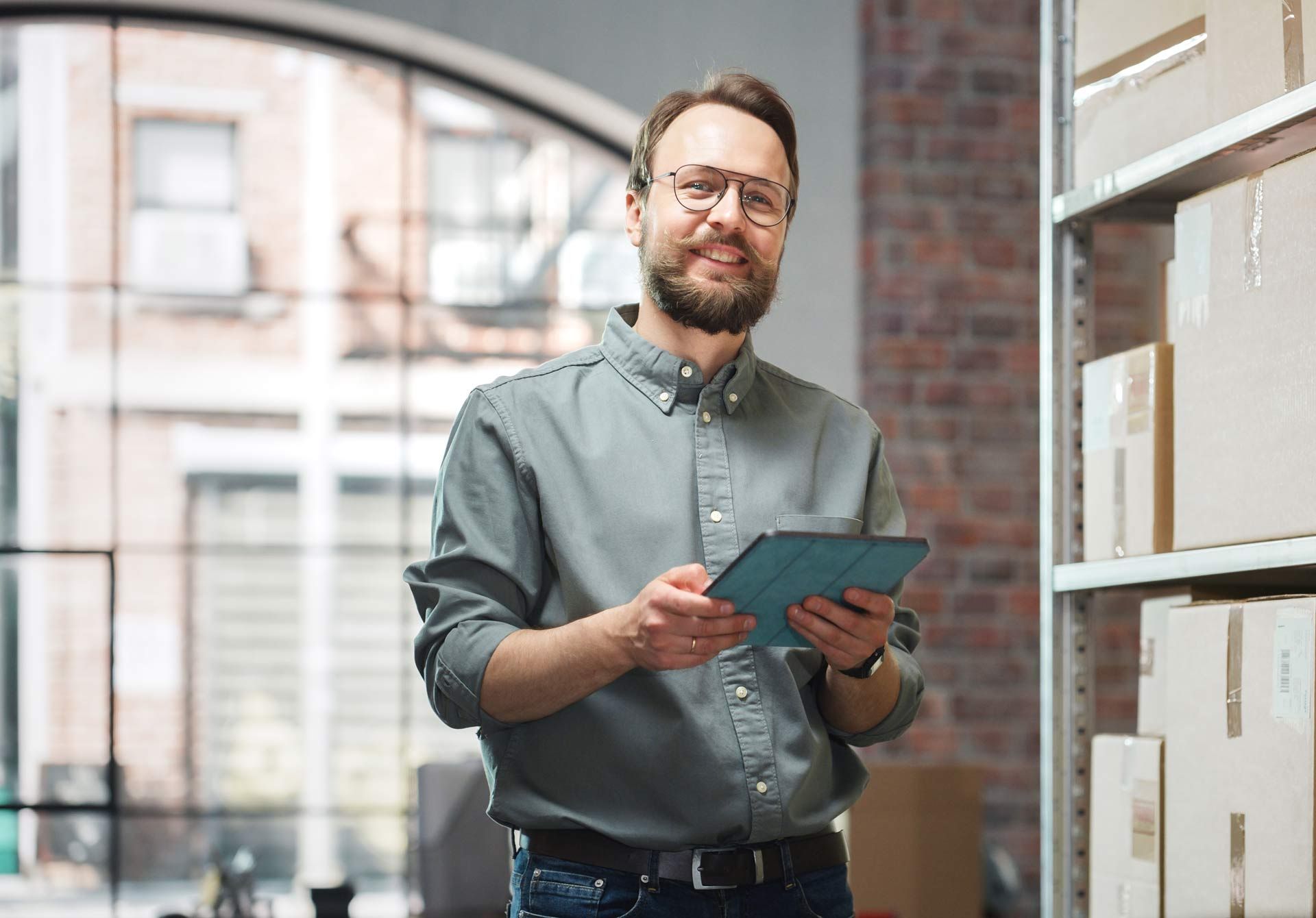 Man in glasses smiling, holding tablet, standing in warehouse, near shelves of boxes.