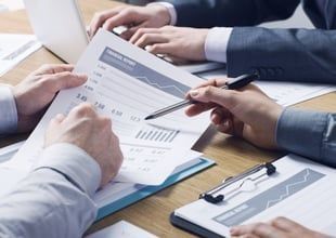 People in suits reviewing financial documents, using a laptop, and pointing with a pen at a chart.