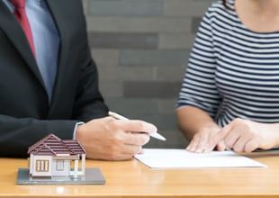 Man in suit and woman at table, signing a document with a model house nearby.