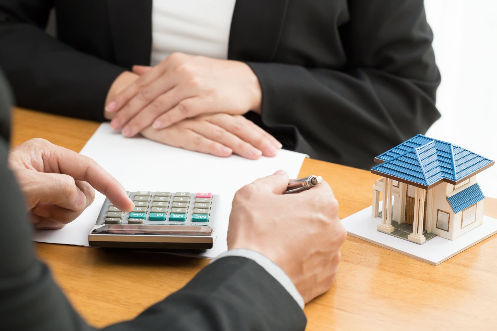 Person using a calculator, with a model house and paperwork on a desk. Another person's hands are visible.