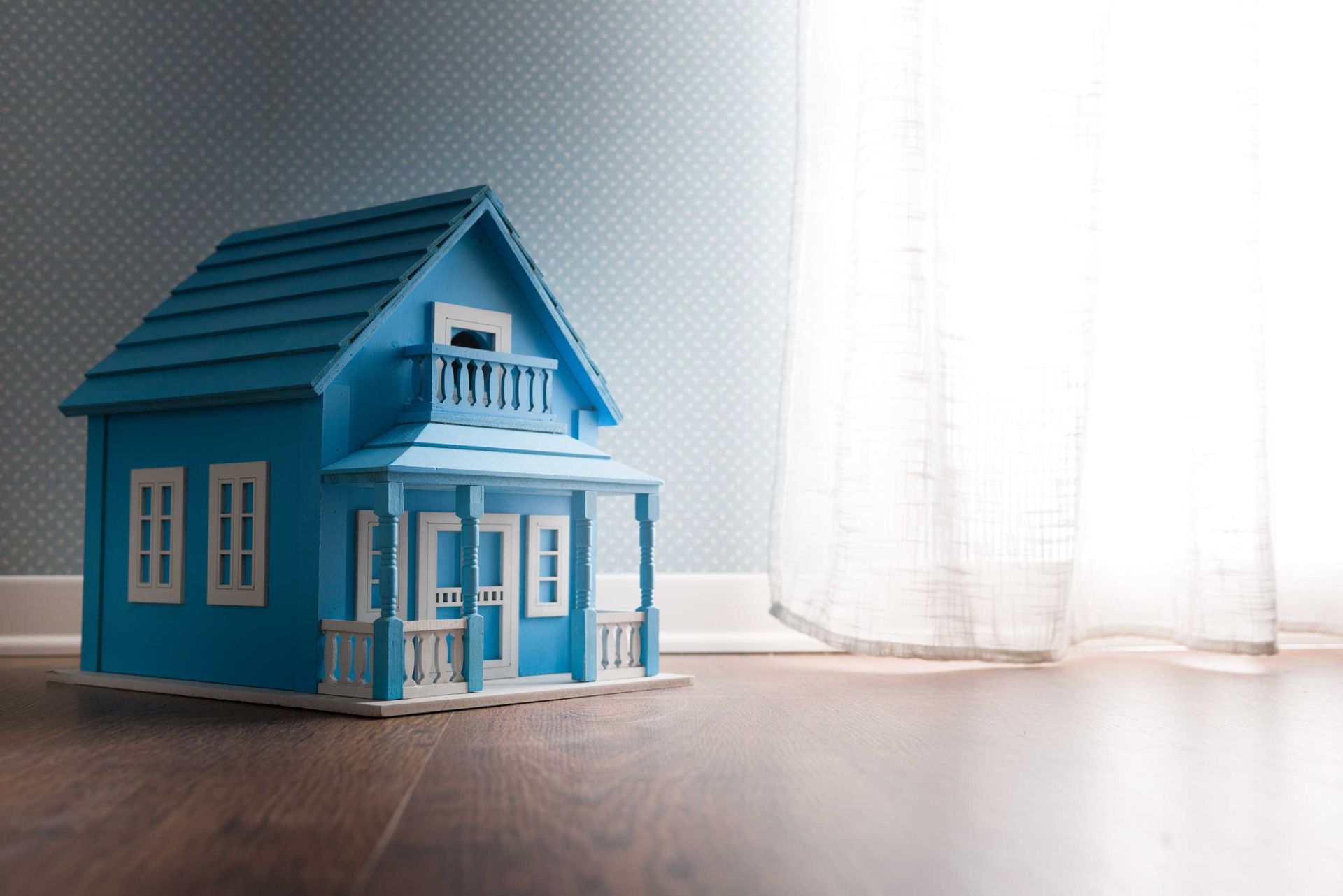 Blue toy house on a wooden floor, near a white curtain, against a blue wall.