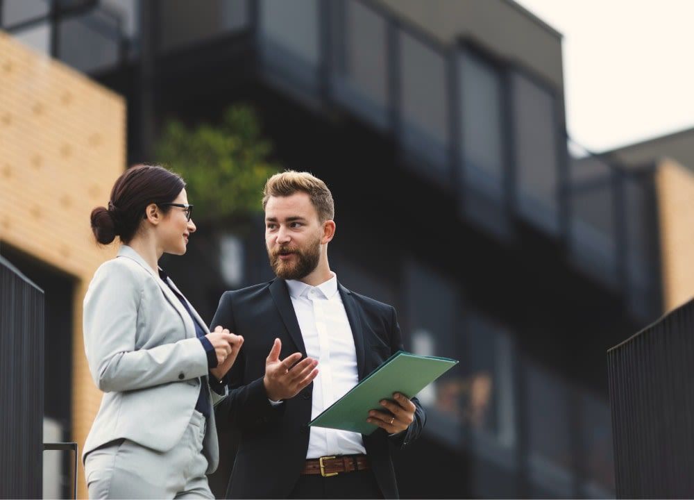 Two professionals in suits talking outside modern building; man holds papers.