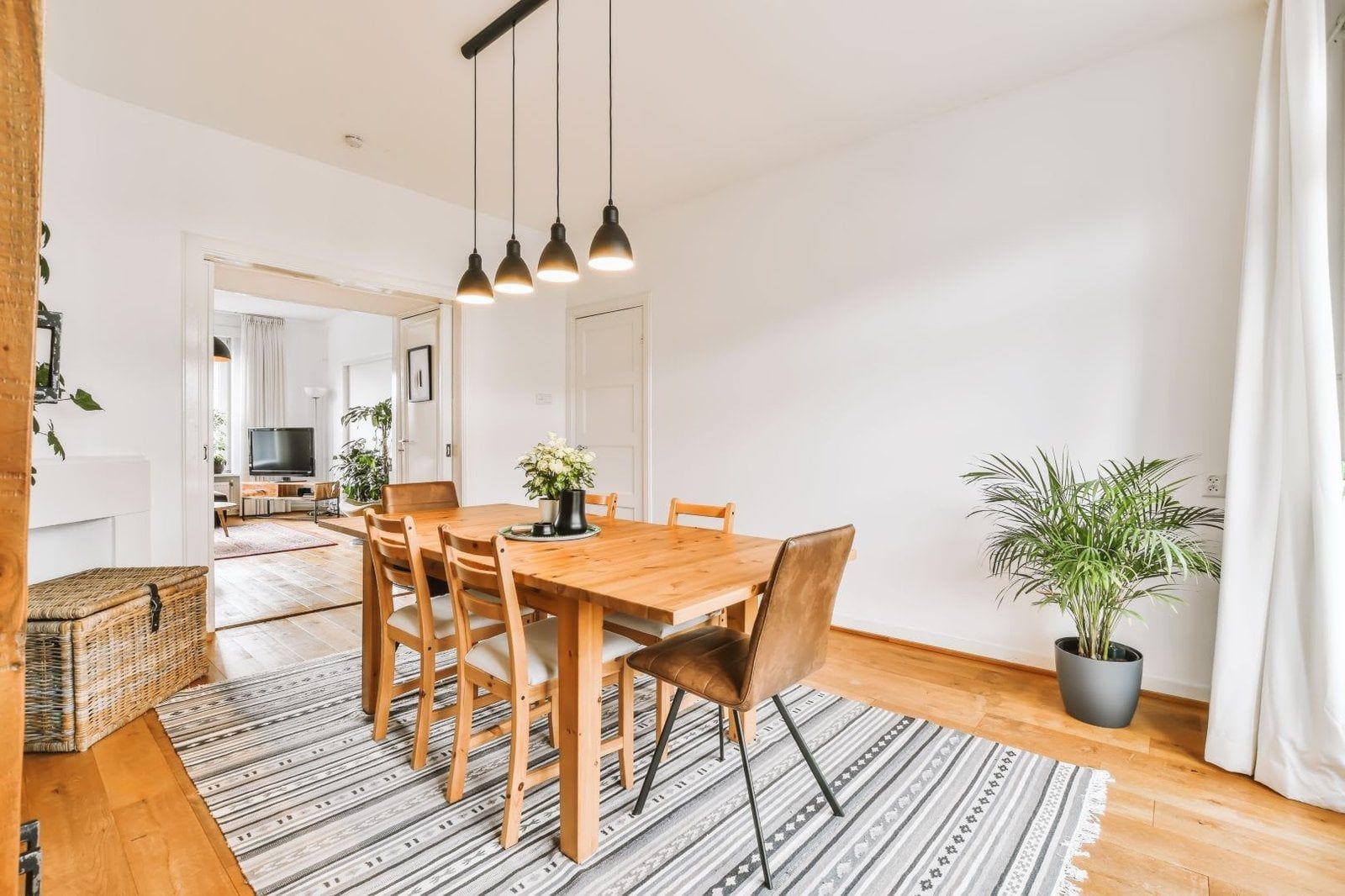 Dining room with wooden table, chairs, rug, and pendant lights. A potted plant sits near a window.
