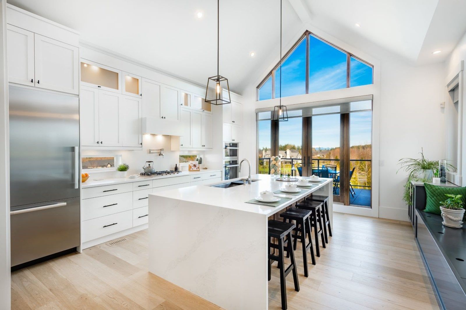 Modern white kitchen with island seating, stainless steel appliances, and large windows with outdoor view.