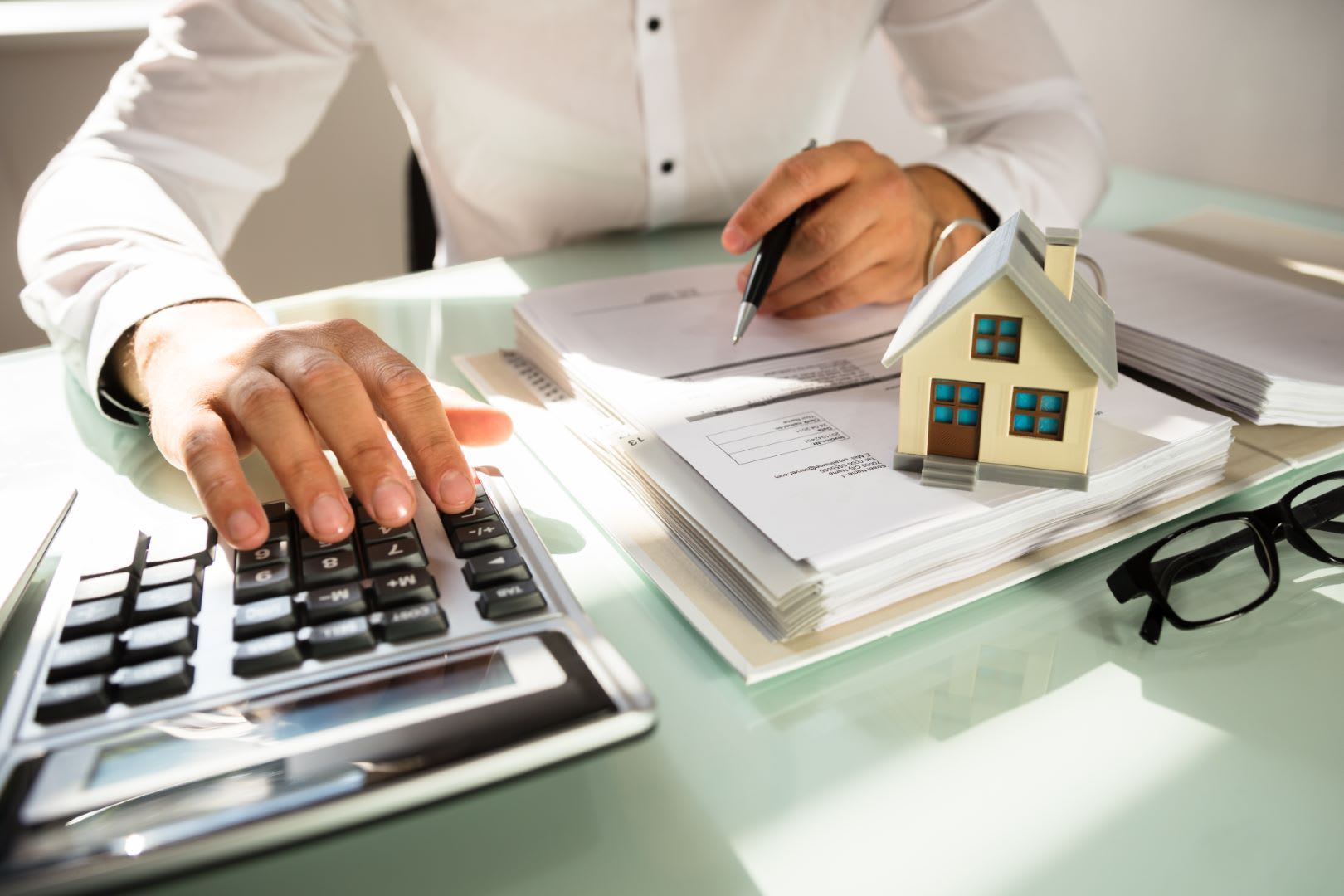 Person using calculator next to miniature house and documents, possibly calculating housing costs.