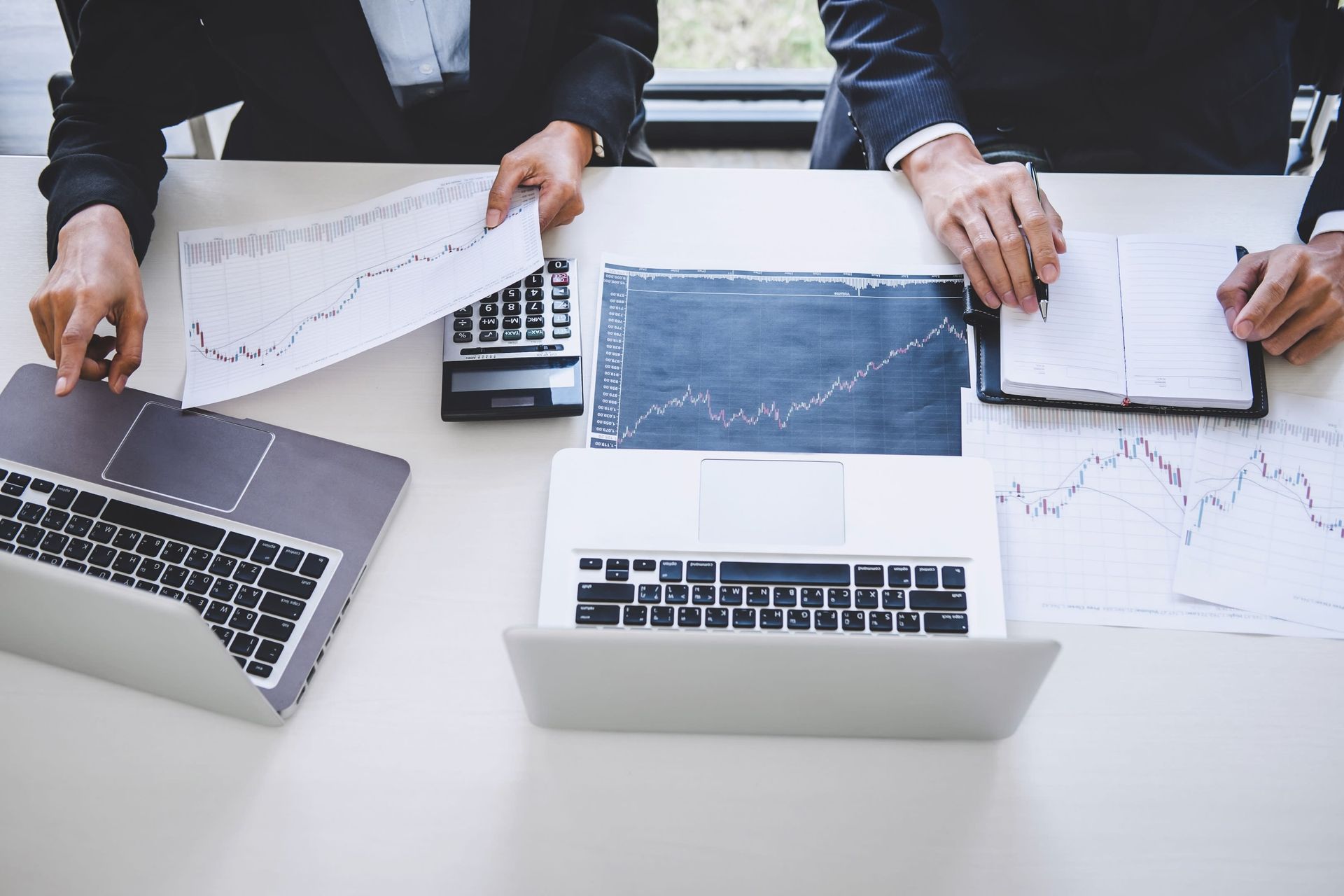 Two people in suits analyzing charts and using laptops at a desk.