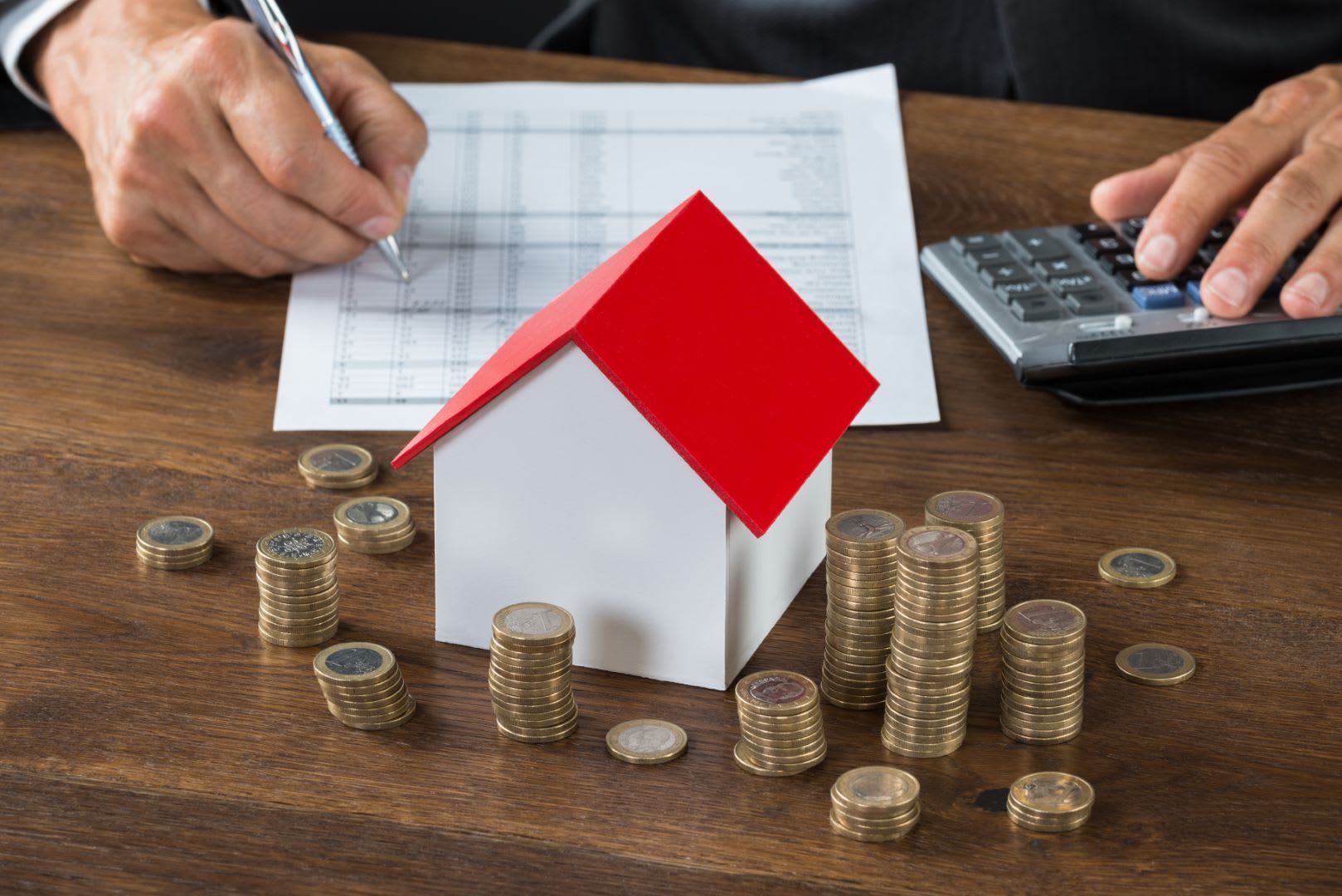Person calculating costs with calculator and paper, with a model house and stacks of coins on a wooden desk.