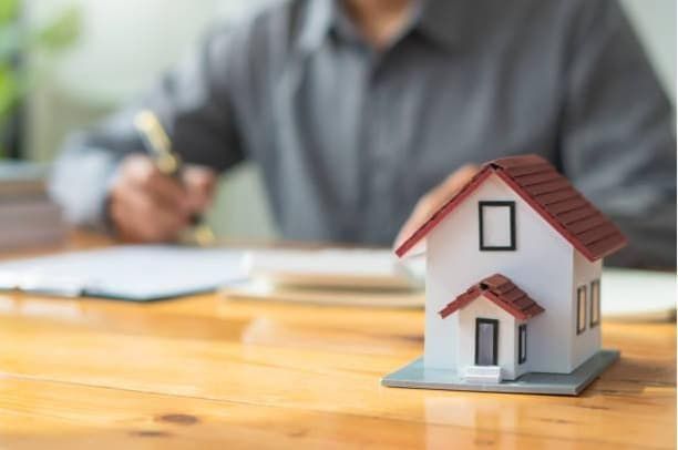 Miniature house on a table; person writing in the background; document and calculator present.