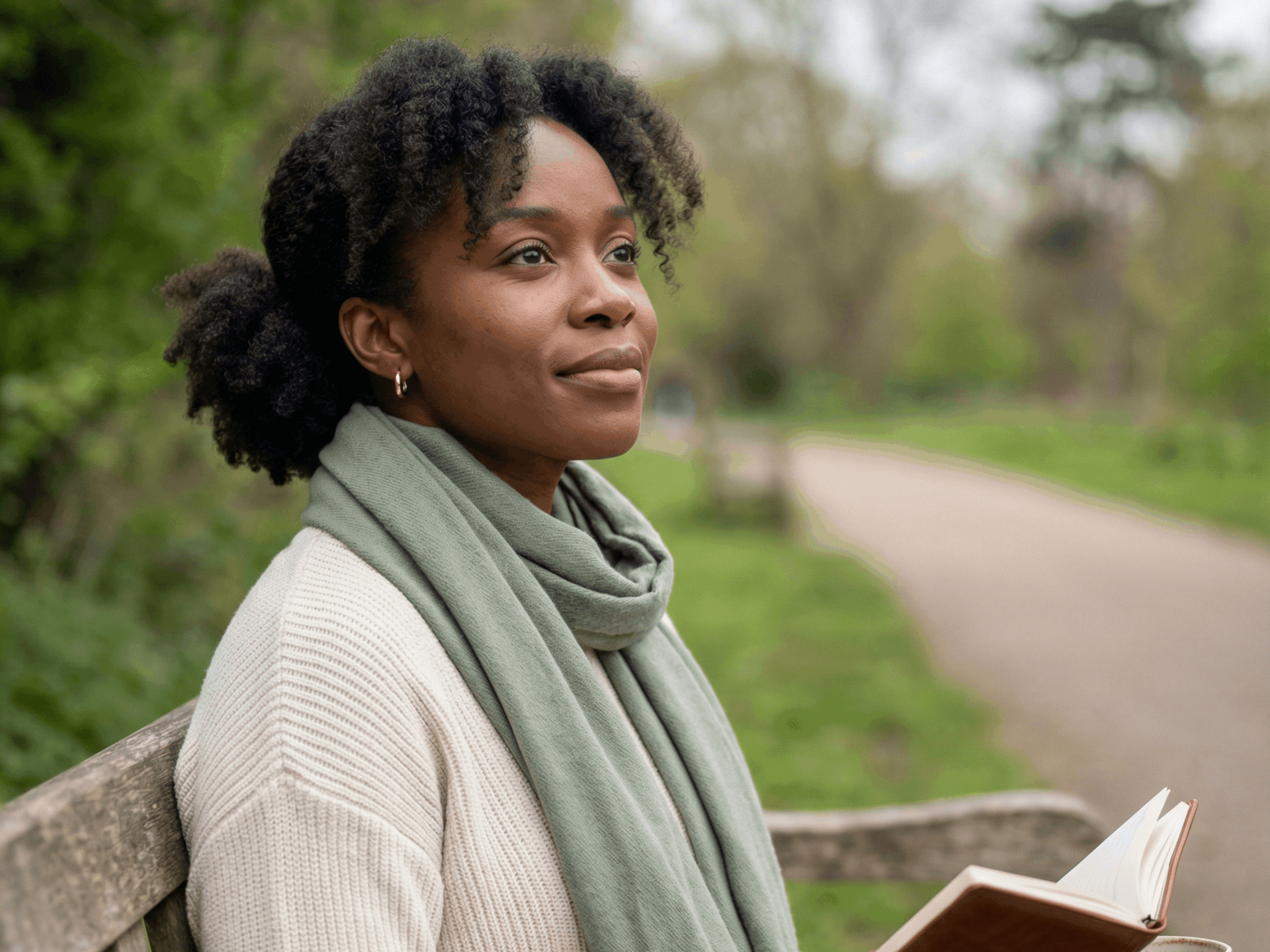 Woman with curly hair sits on a bench, looking thoughtfully to the side. Wearing scarf and sweater, holding a book in a park.
