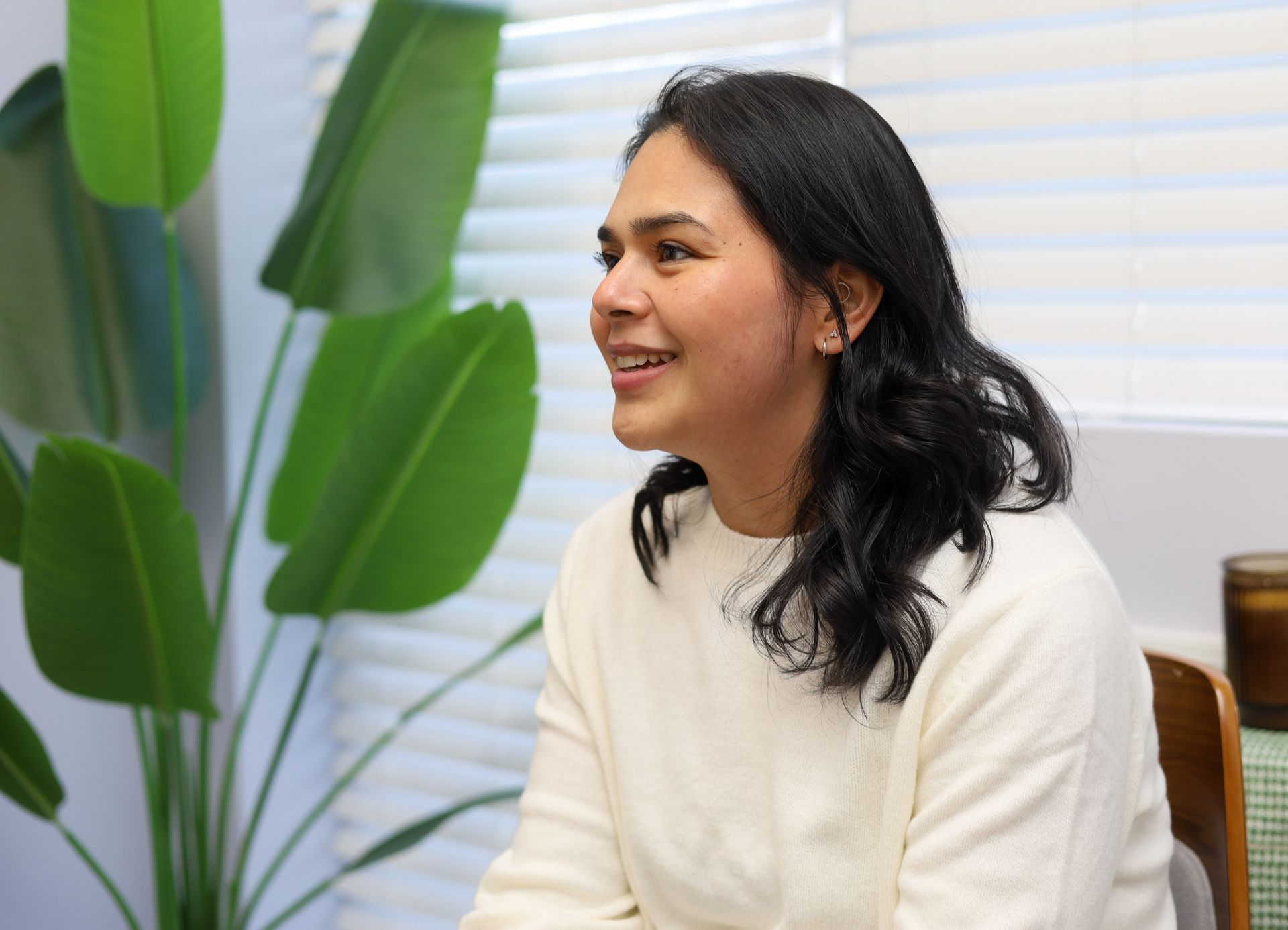 Woman in a white sweater smiles, seated indoors, near a large green plant.