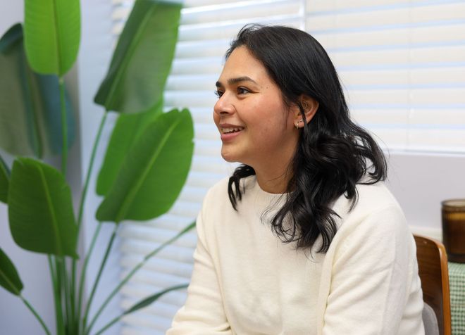 Woman in a white sweater smiles, seated indoors, near a large green plant.