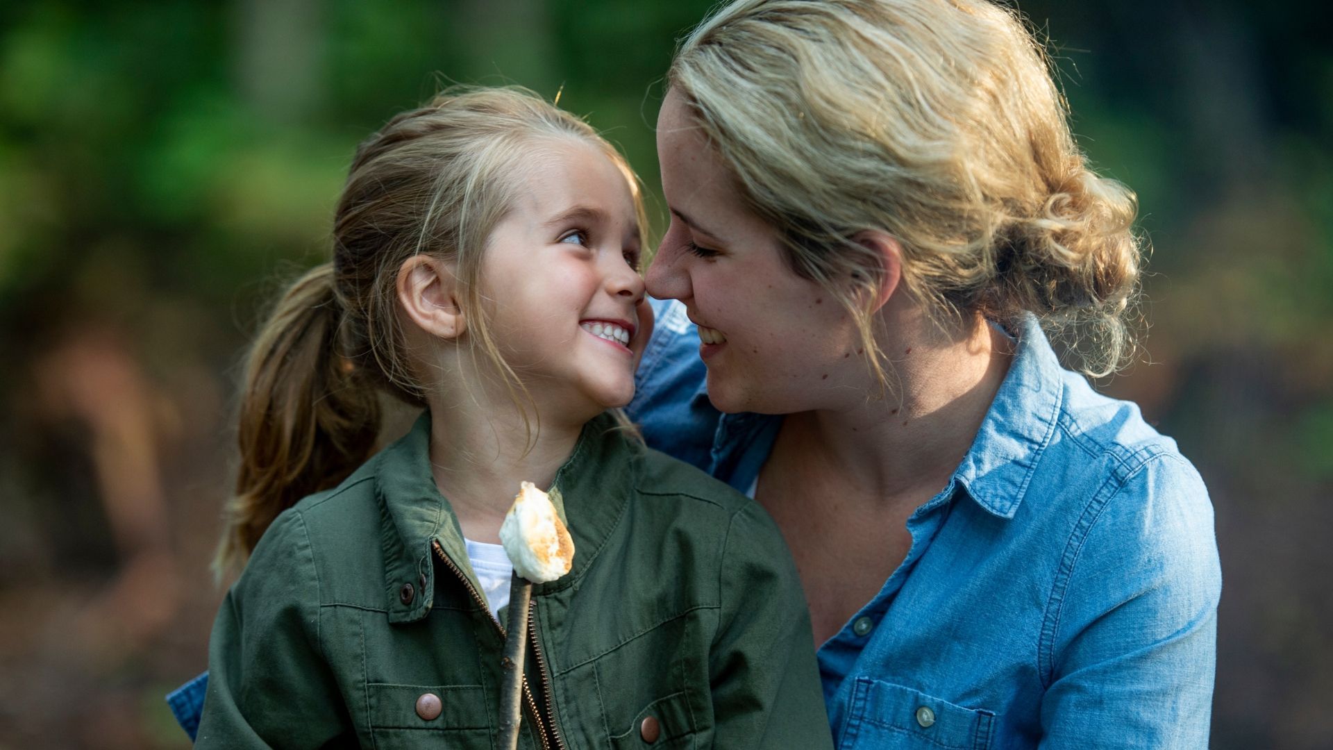 Woman and child smiling at each other, holding a marshmallow on a stick outdoors.