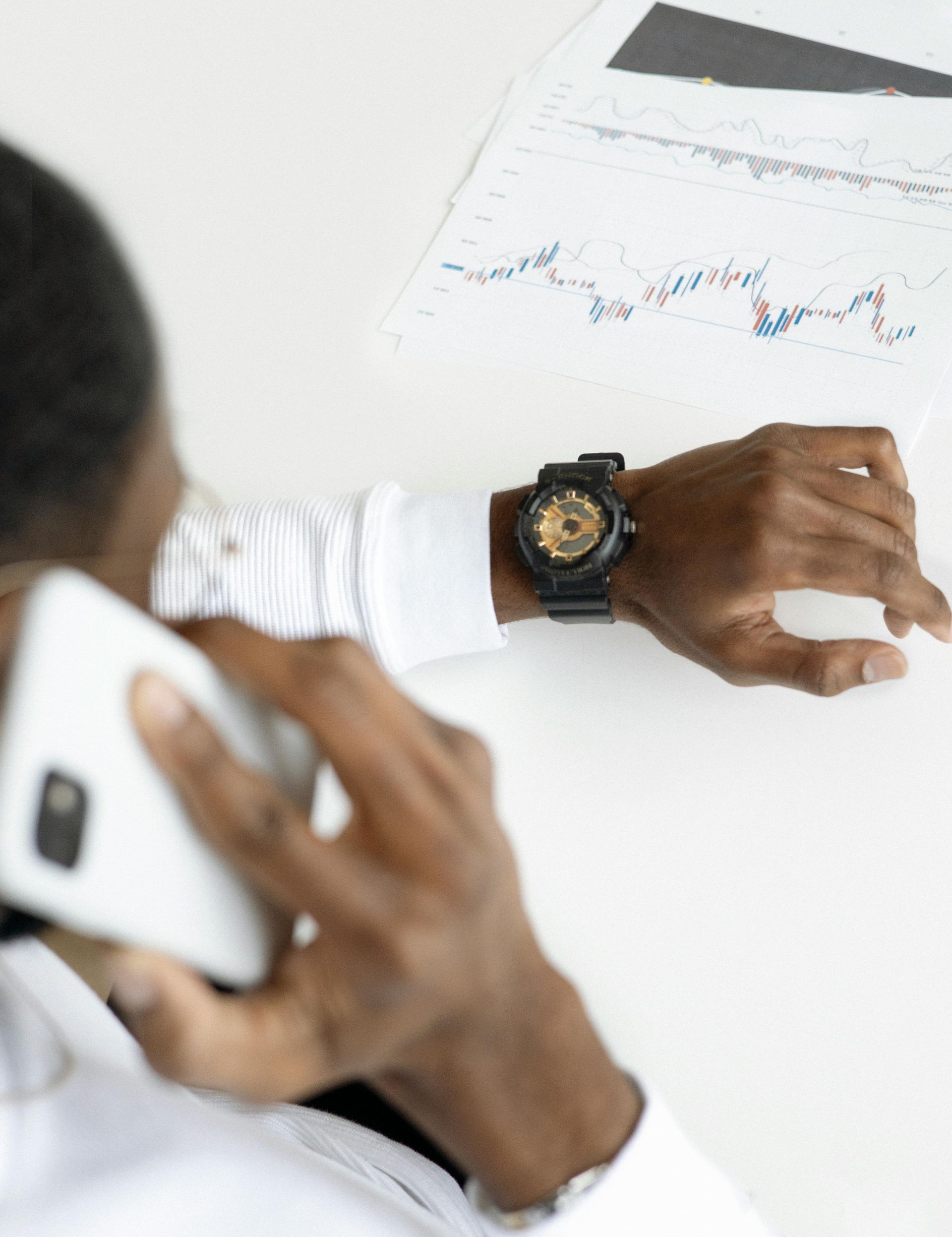 Person on phone, looking at watch, next to financial graph on white table.