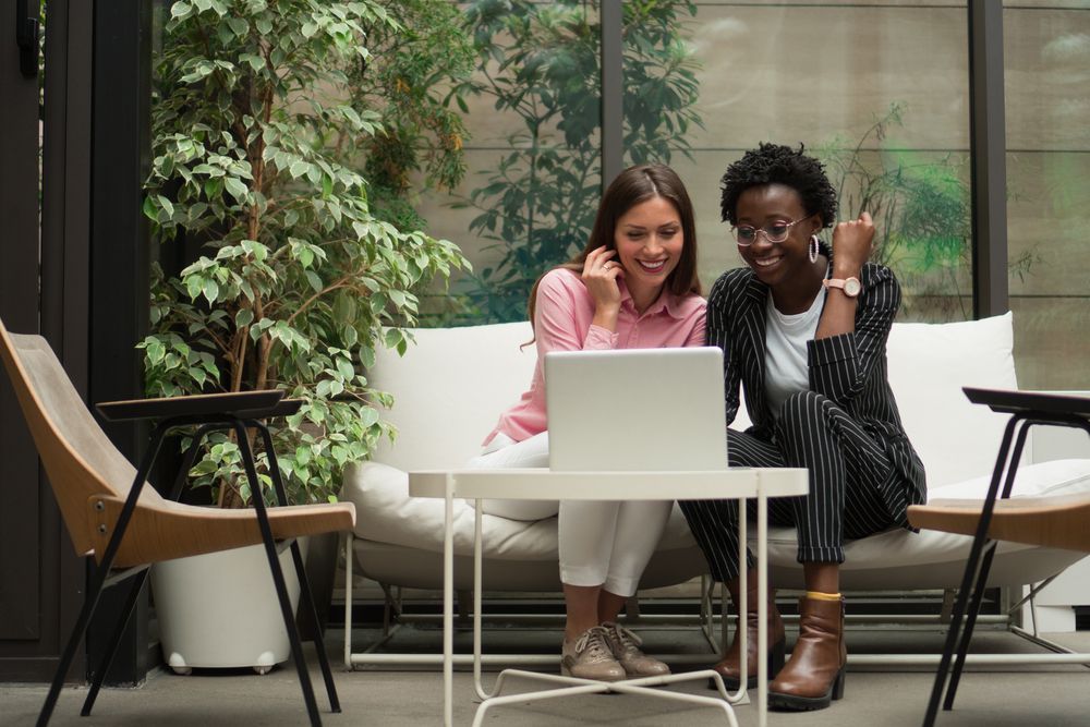 two women are sitting on a couch looking at a laptop with greenery around them.