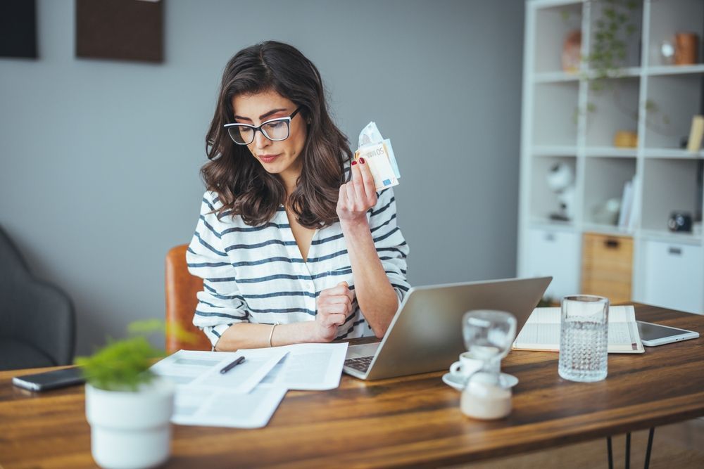 A woman is sitting at a desk with a laptop and holding a piece of paper.