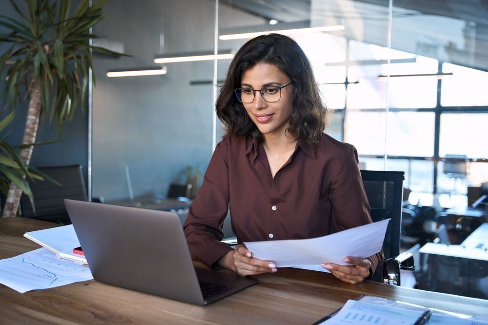 A woman is sitting at a desk with a laptop and a piece of paper.