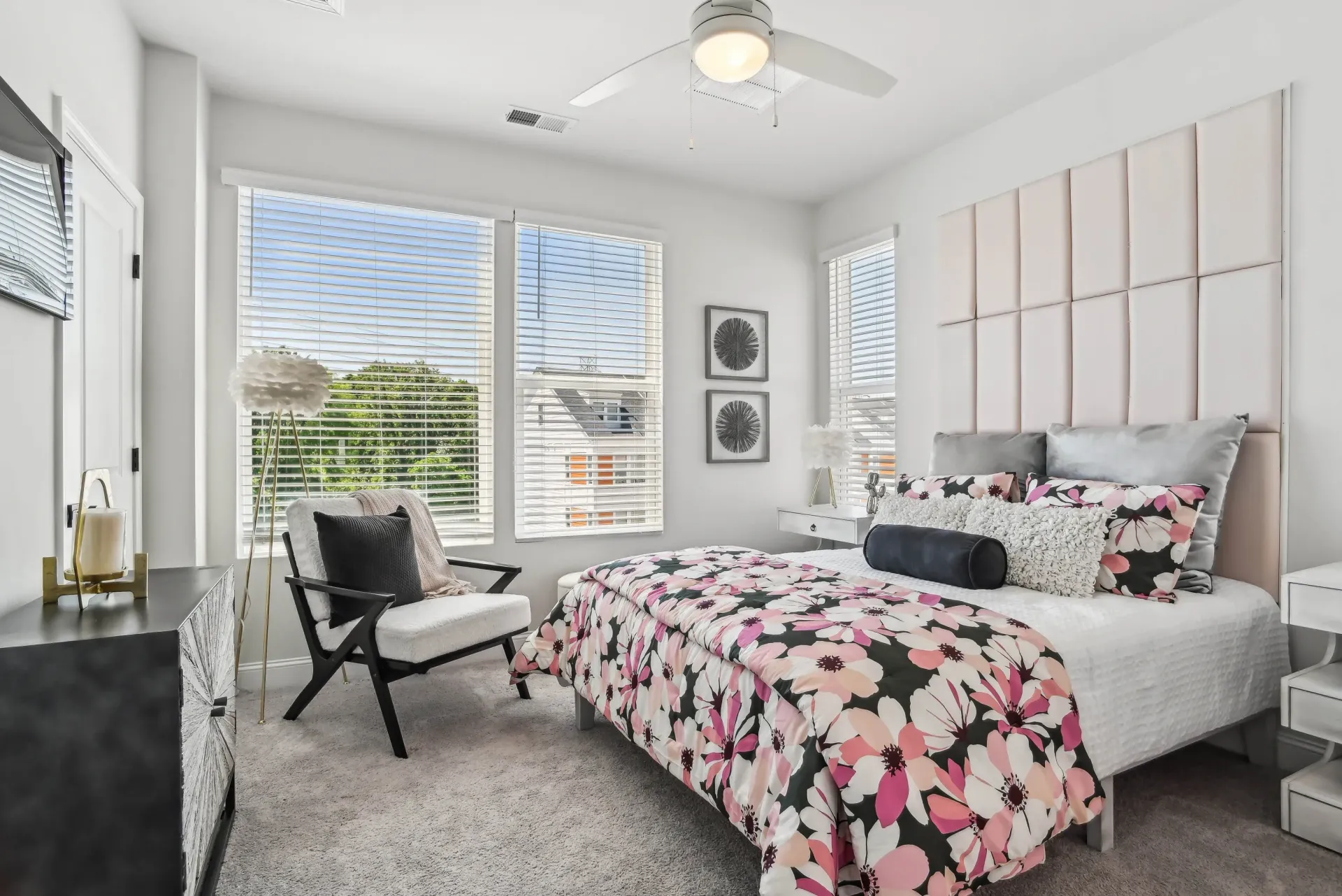 Bedroom in a modern apartment with a tall padded headboard, floral bedding, and large windows.