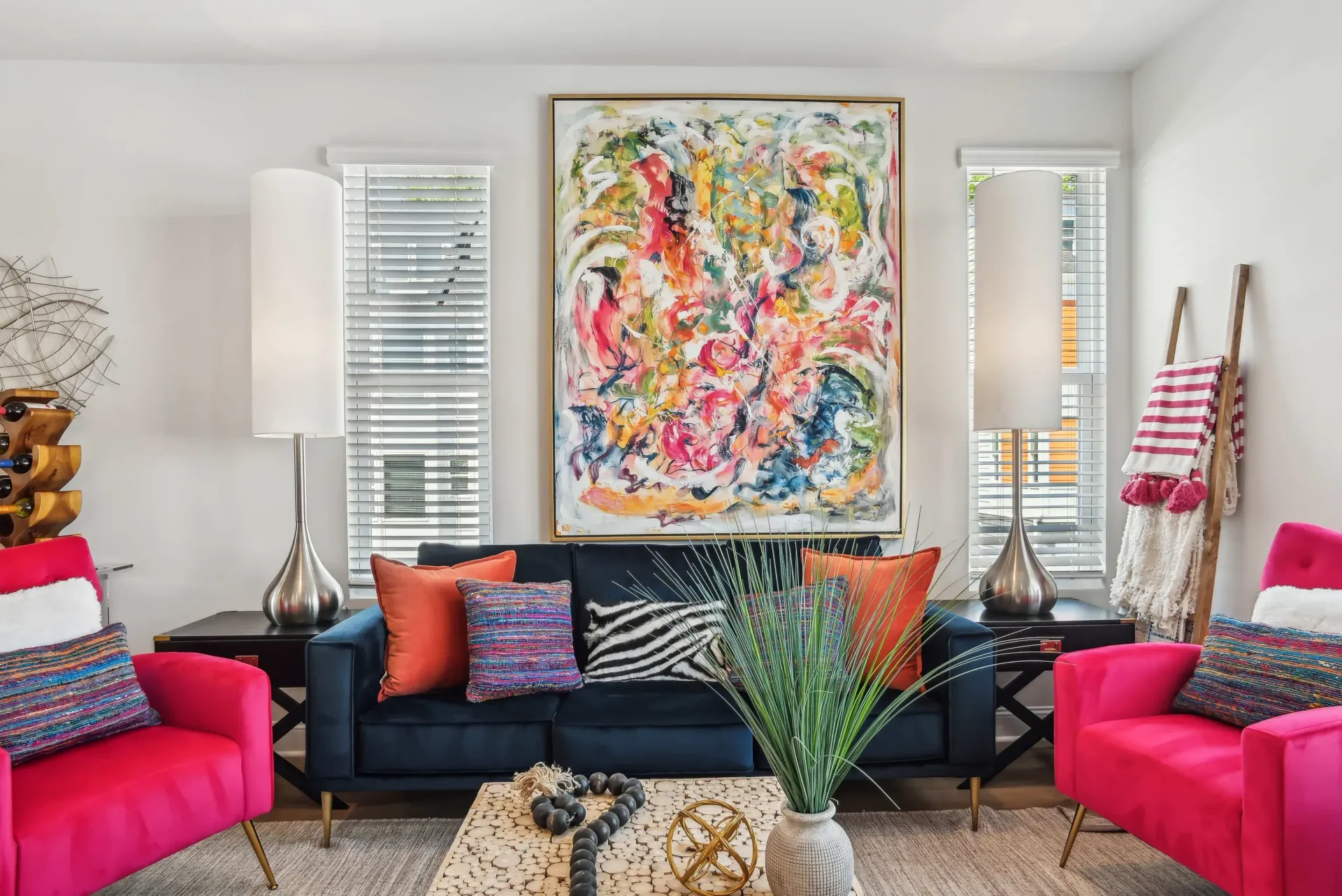 Living room interior with a navy sofa, pink accent chairs, abstract wall art, and two tall lamps.