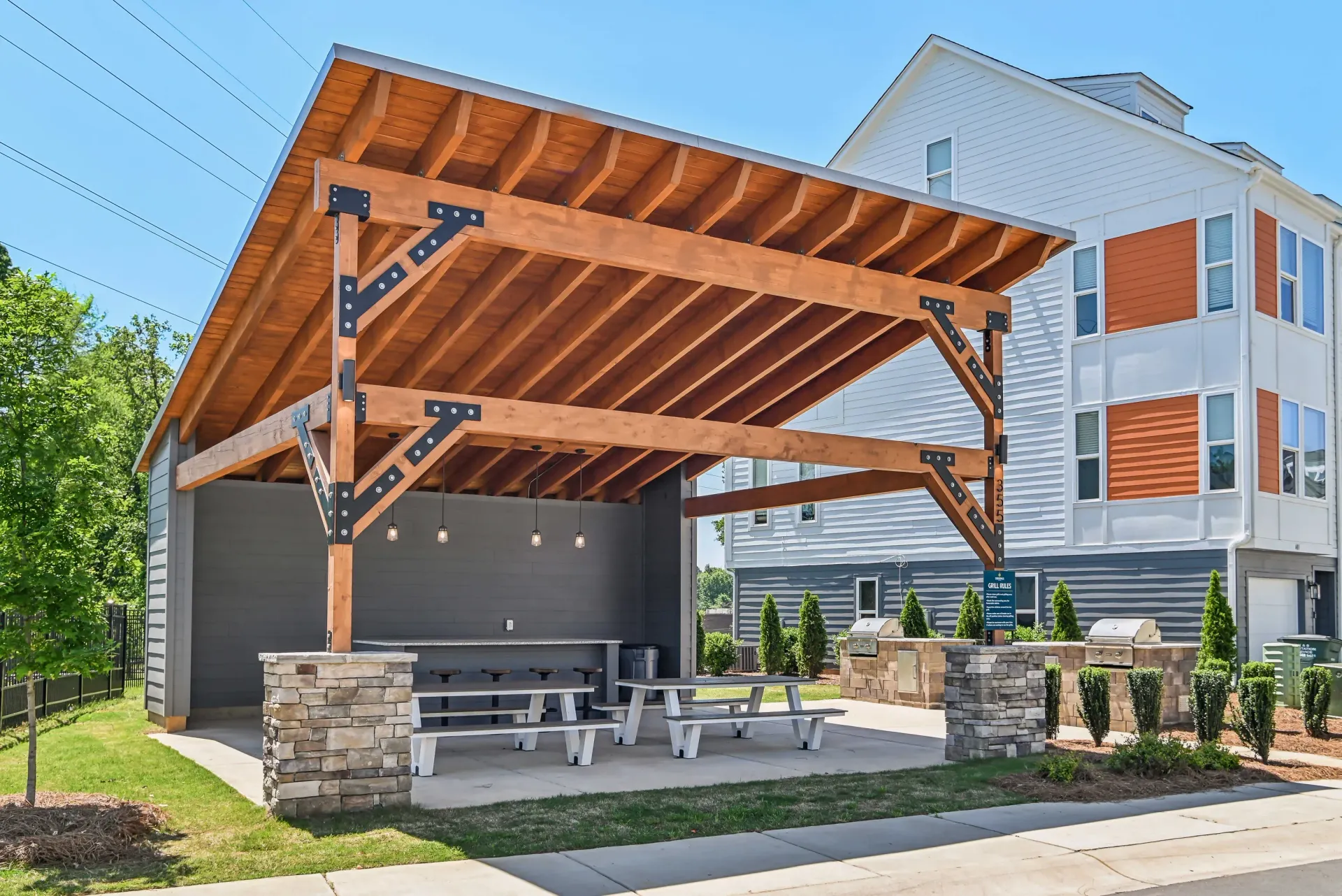 Outdoor communal grilling area under a wooden pergola with stone pillars, grills, and picnic tables.